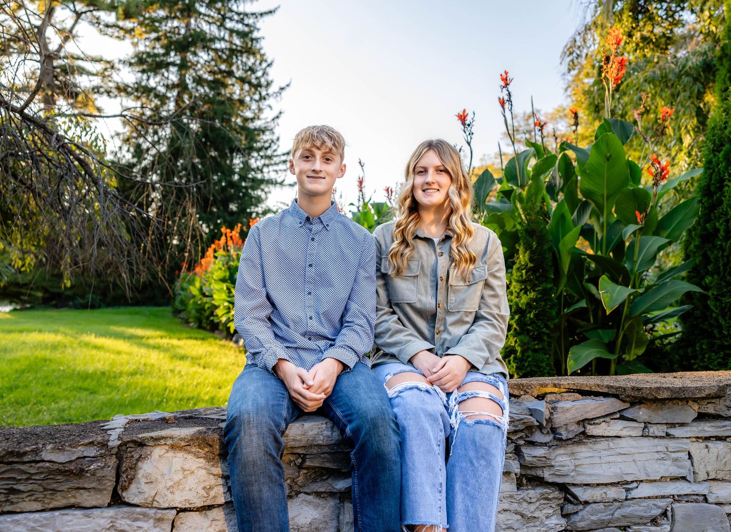 Two people sit on a stone wall with red flowers and greenery in the background.