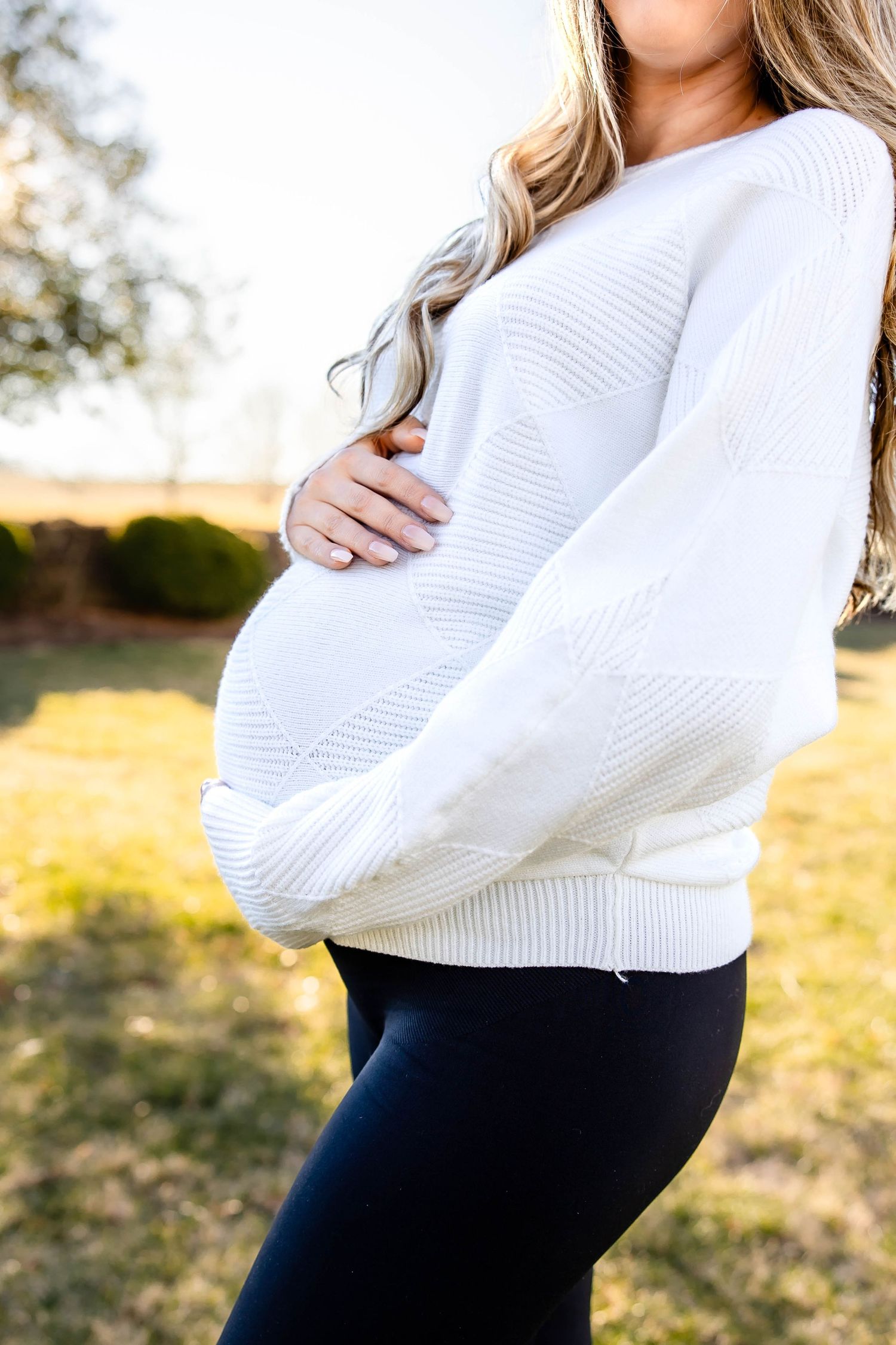 Close-up side view of pregnant belly in textured white sweater against outdoor setting.
