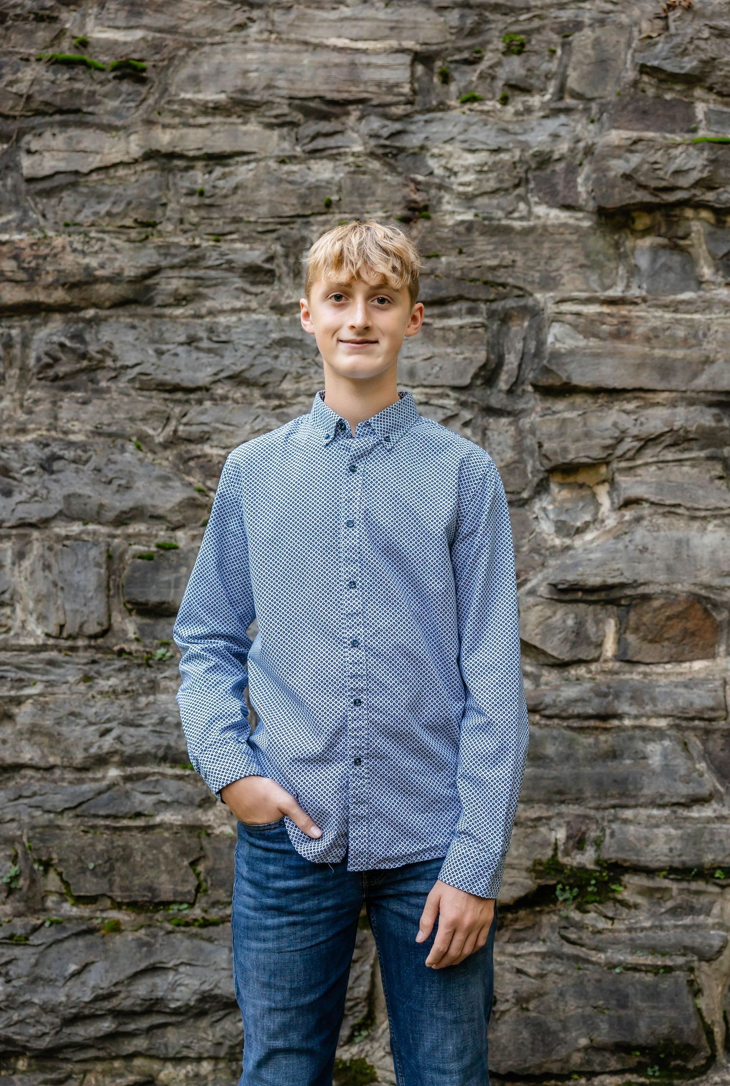 A teenager in a blue button-down shirt poses against a rocky wall background.