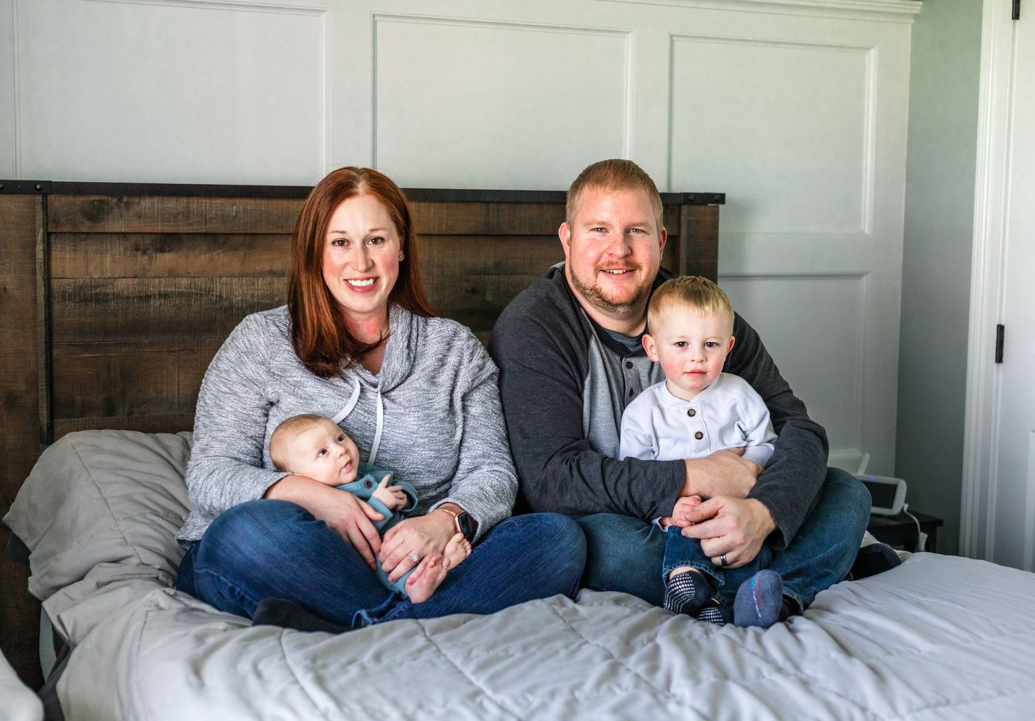 Family sitting together on bed with rustic wooden headboard and grey bedding.