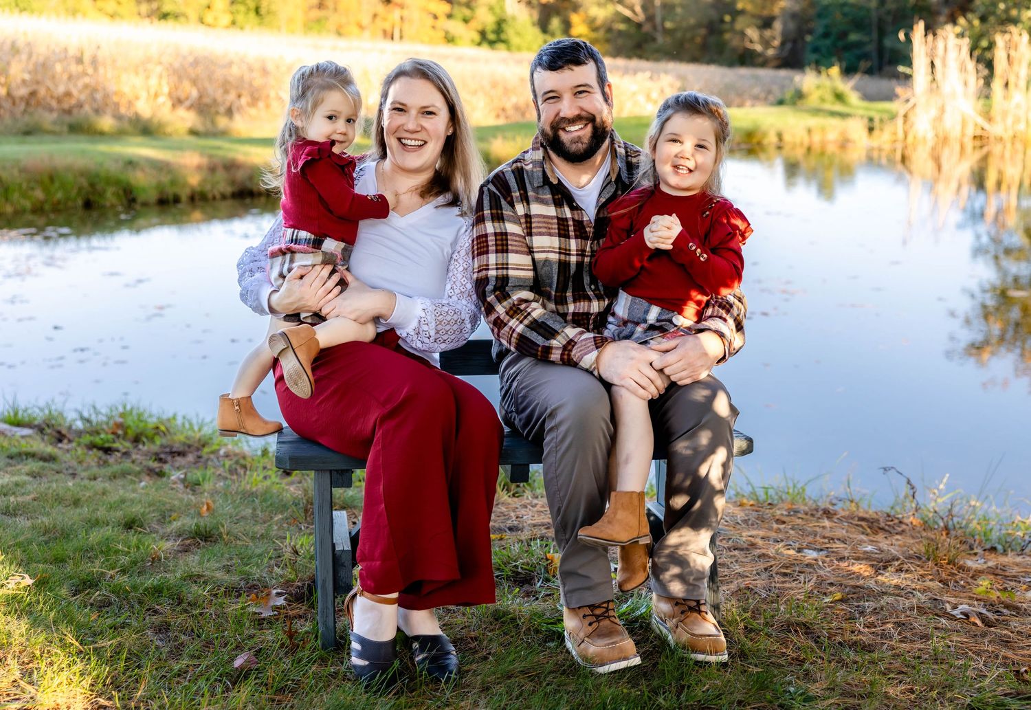 Family of four in coordinating red outfits sits together on bench by scenic pond.