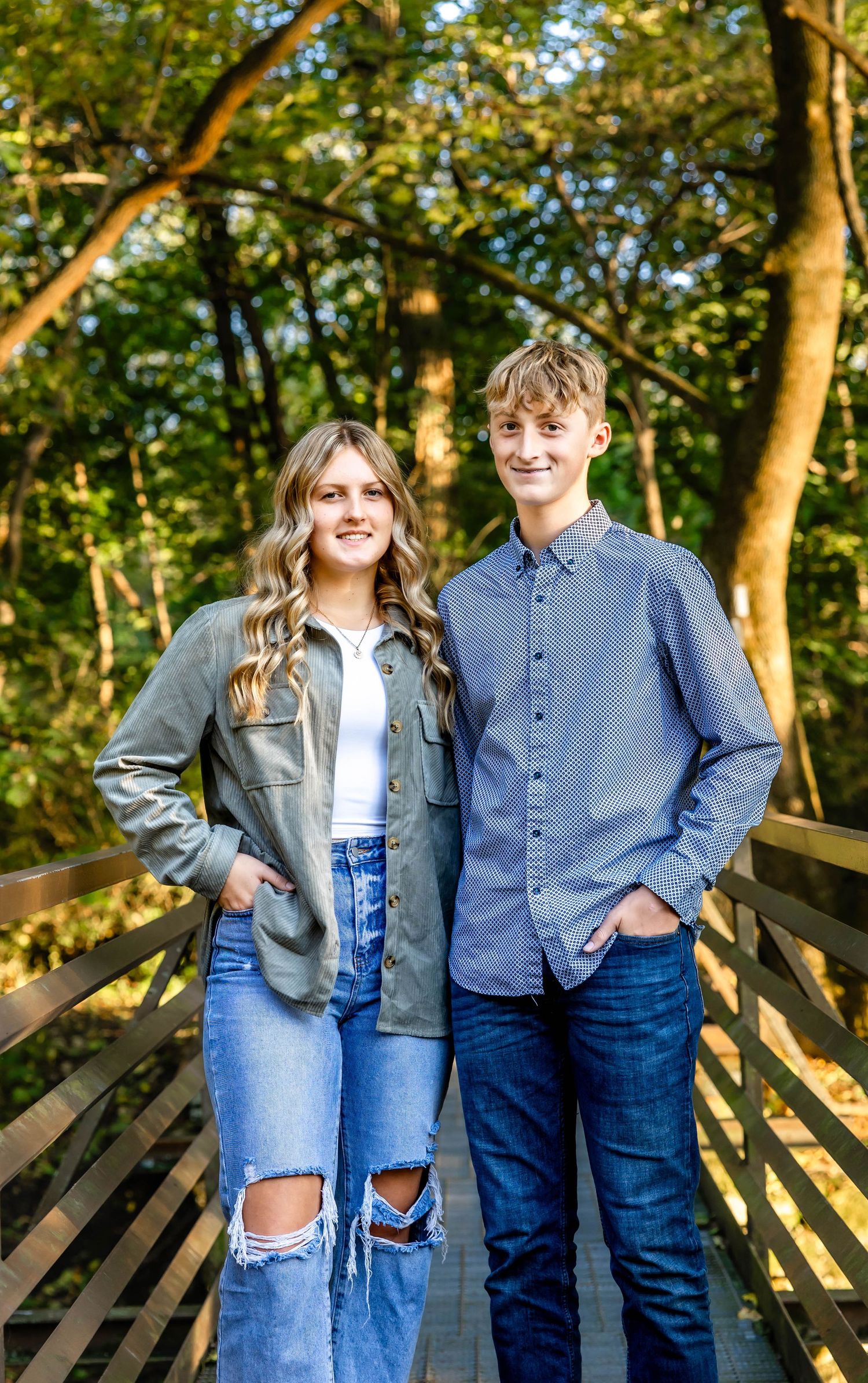 Young couple in casual denim outfits pose together on a wooden boardwalk surrounded by autumn trees.