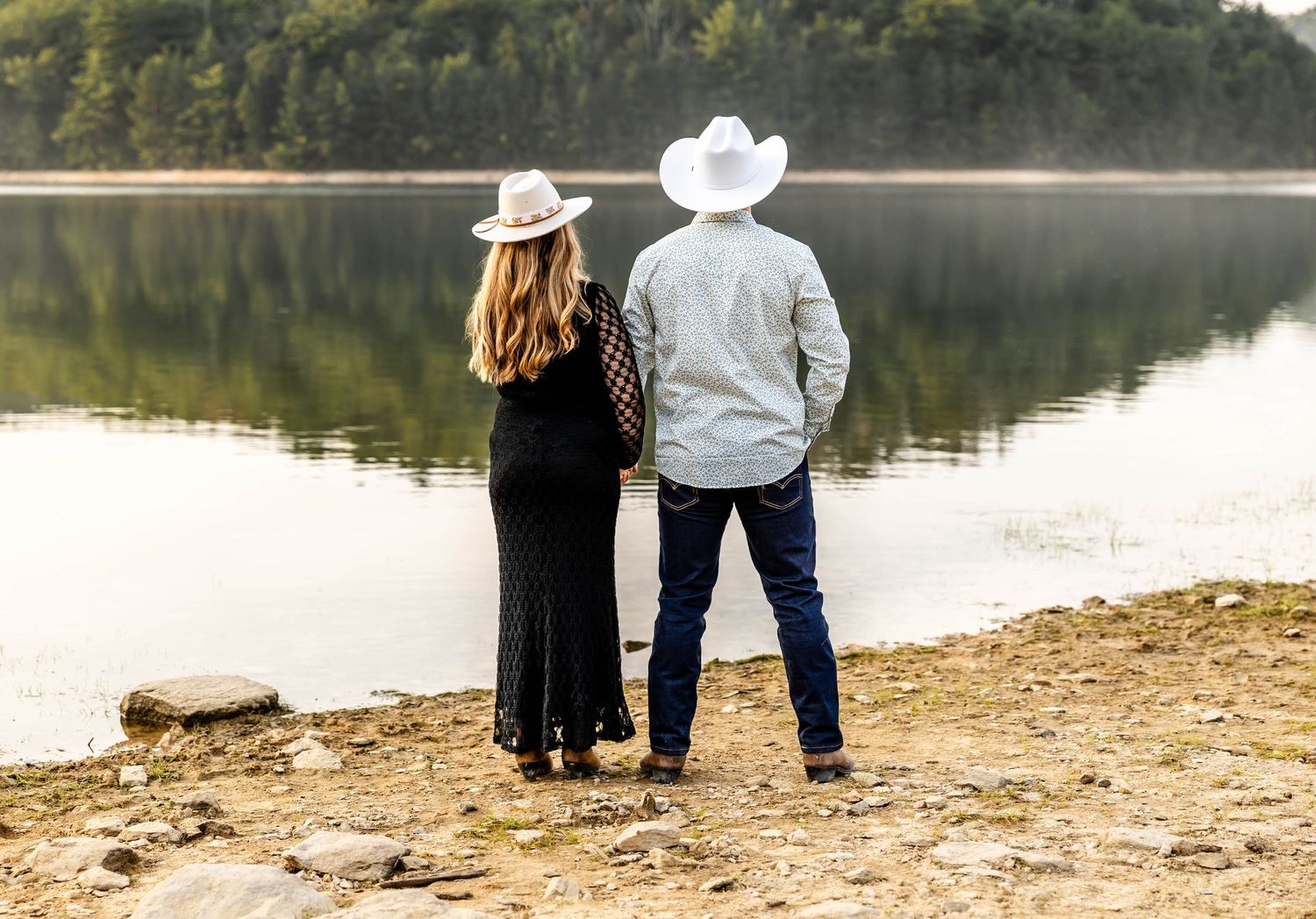 Couple in western attire standing together on lakeside beach at sunset.