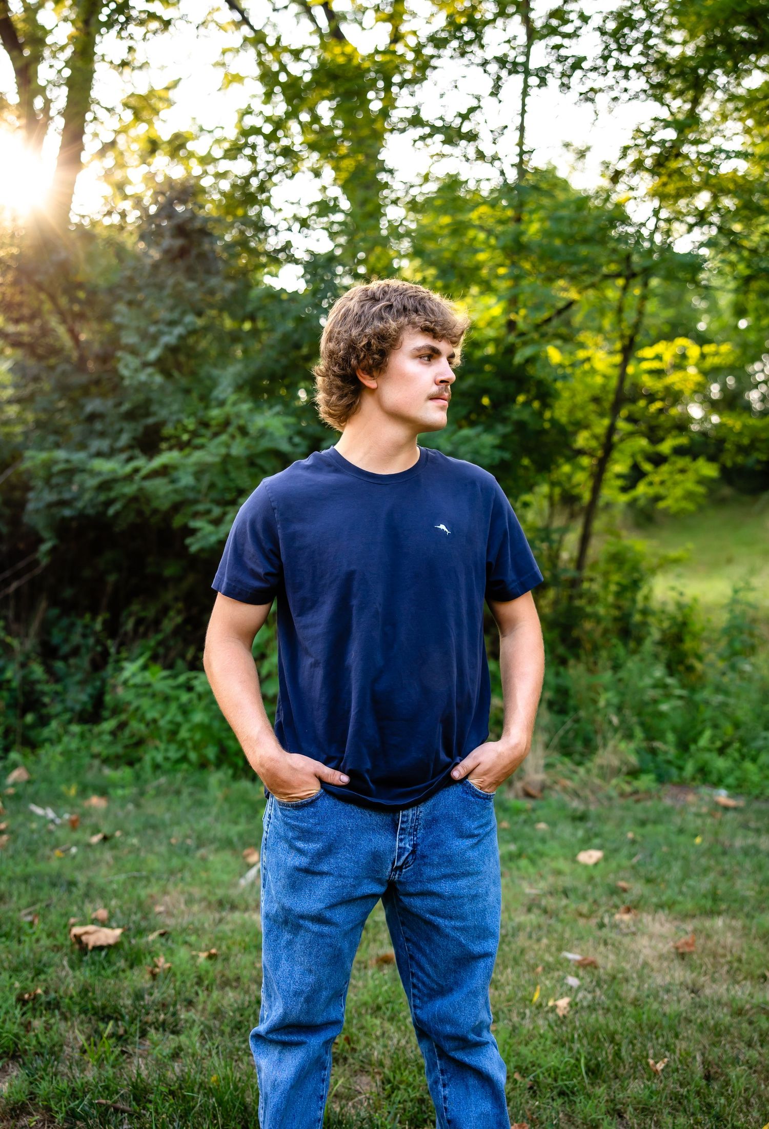 Young person in navy t-shirt and jeans stands in scenic outdoor setting with sunlight peeking through trees.