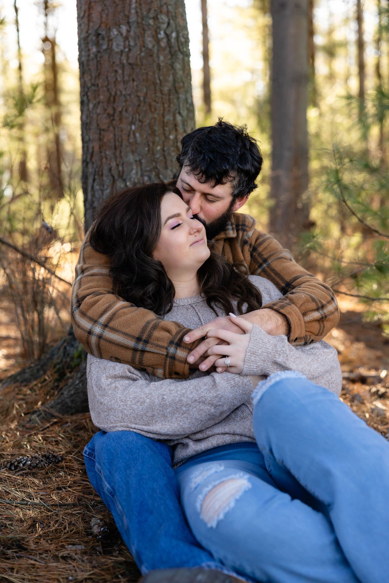 A couple relaxes against a tree trunk in a serene forest setting during golden hour.