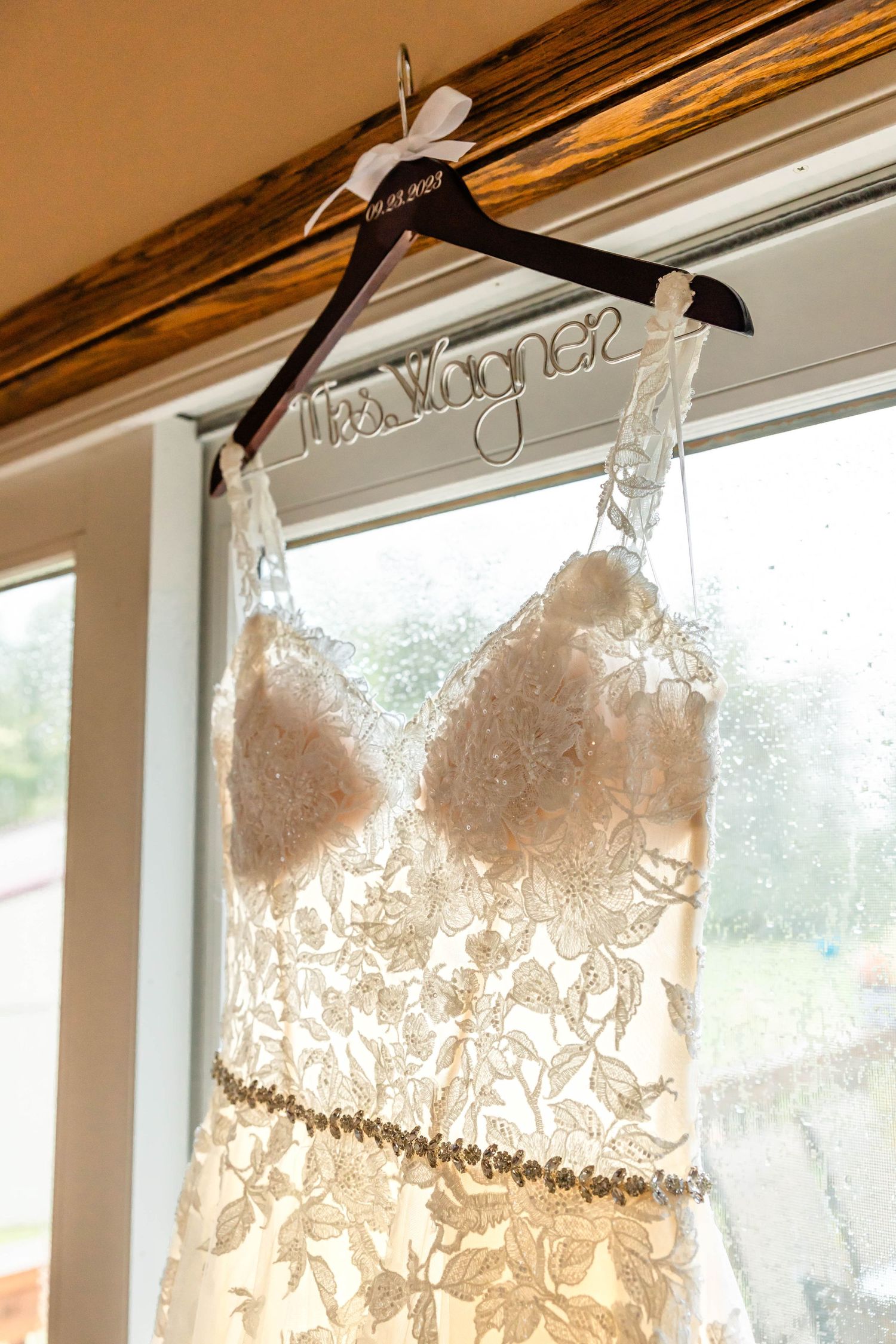 Elegant white lace wedding dress with sweetheart neckline displayed on custom name hanger against window.