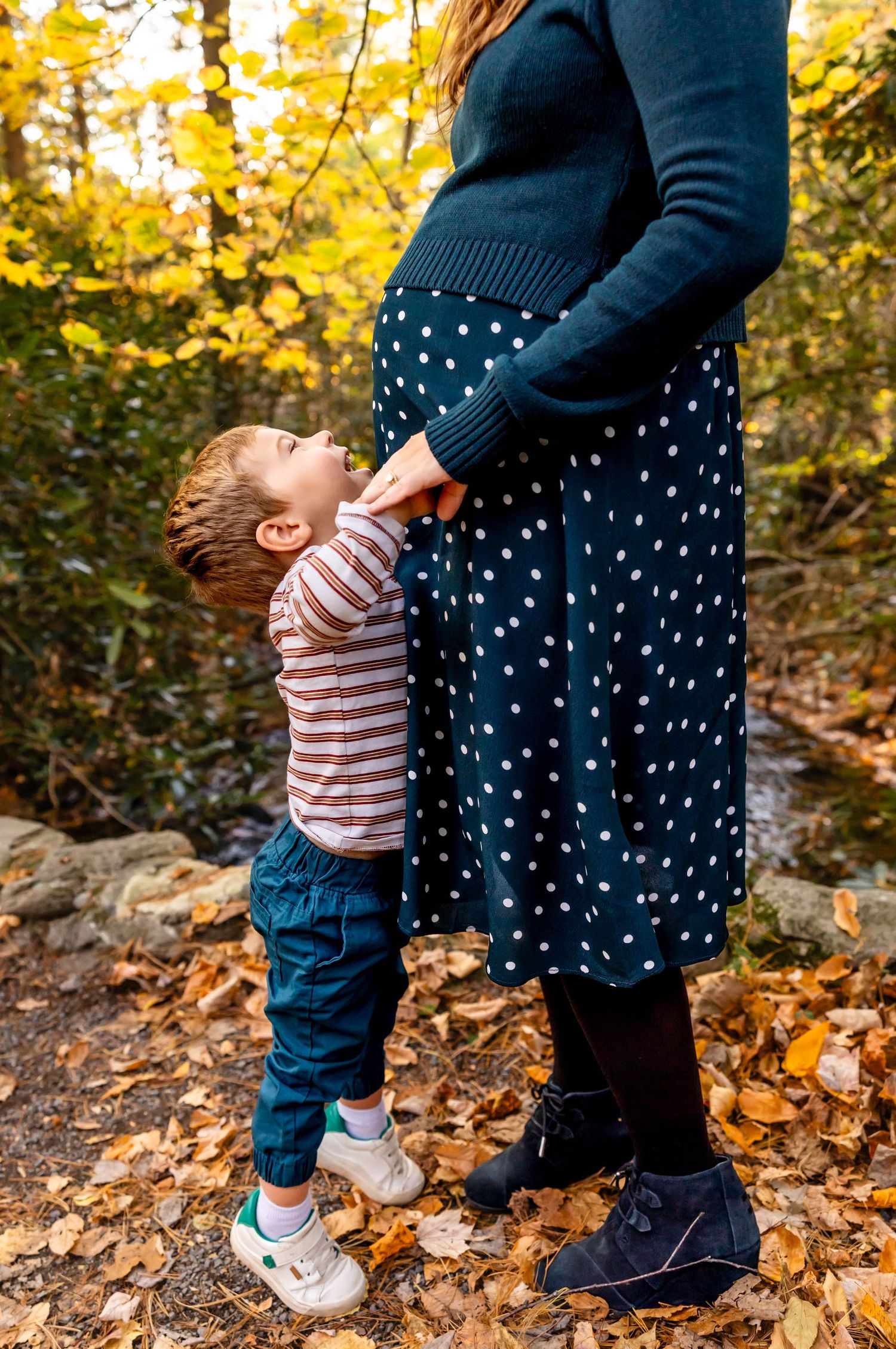 A child in striped shirt reaches up to parent wearing polka dot dress amid fall leaves.