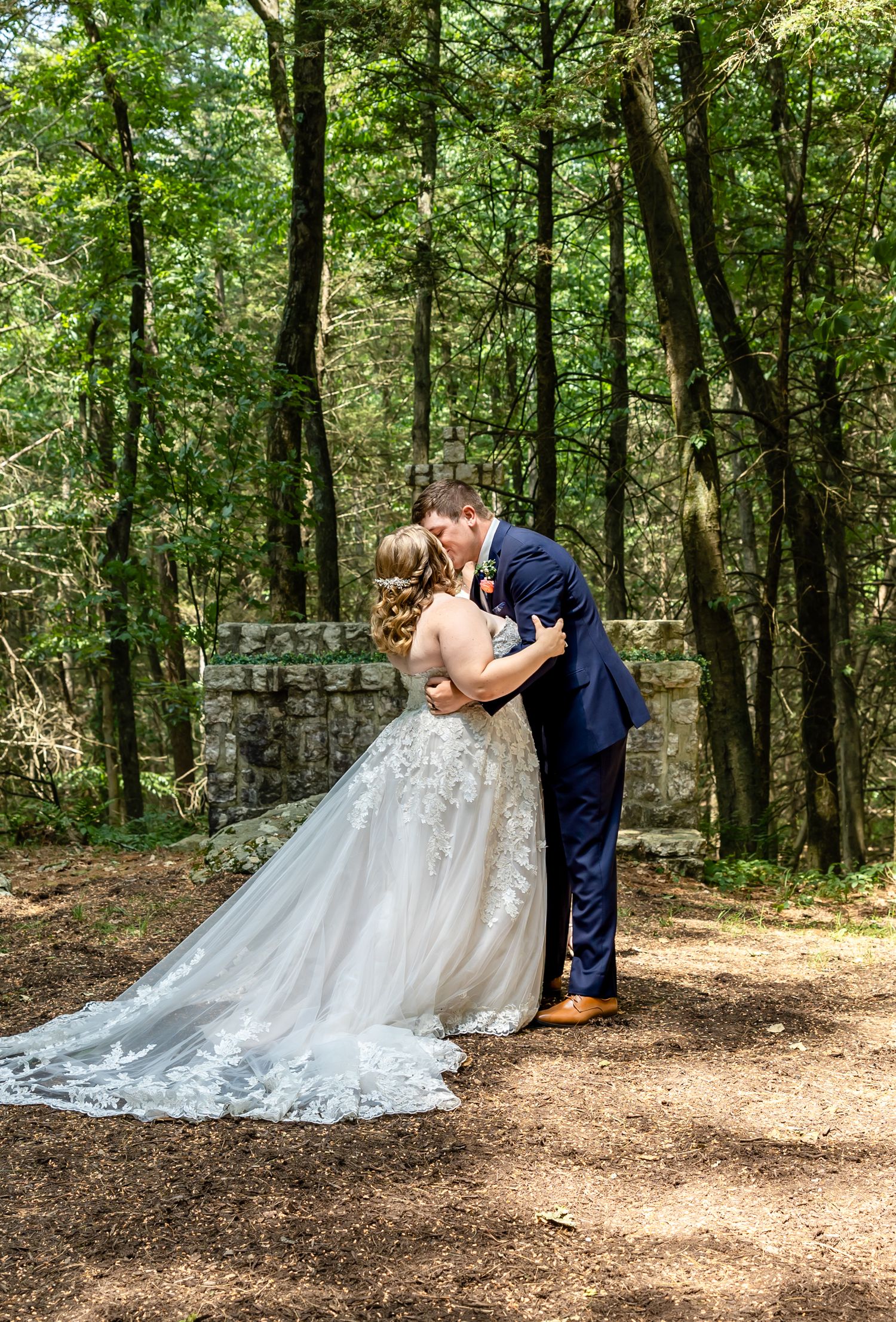Wedding couple sharing romantic kiss on forest path with bride in flowing lace gown.
