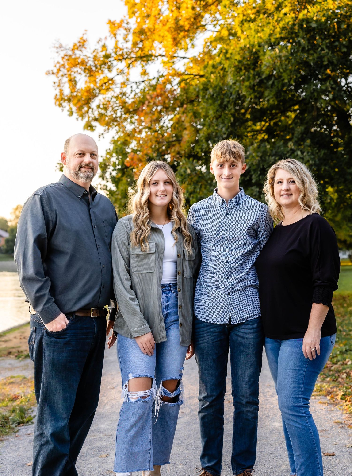 A family of four poses on a path with autumn trees and water in the background.