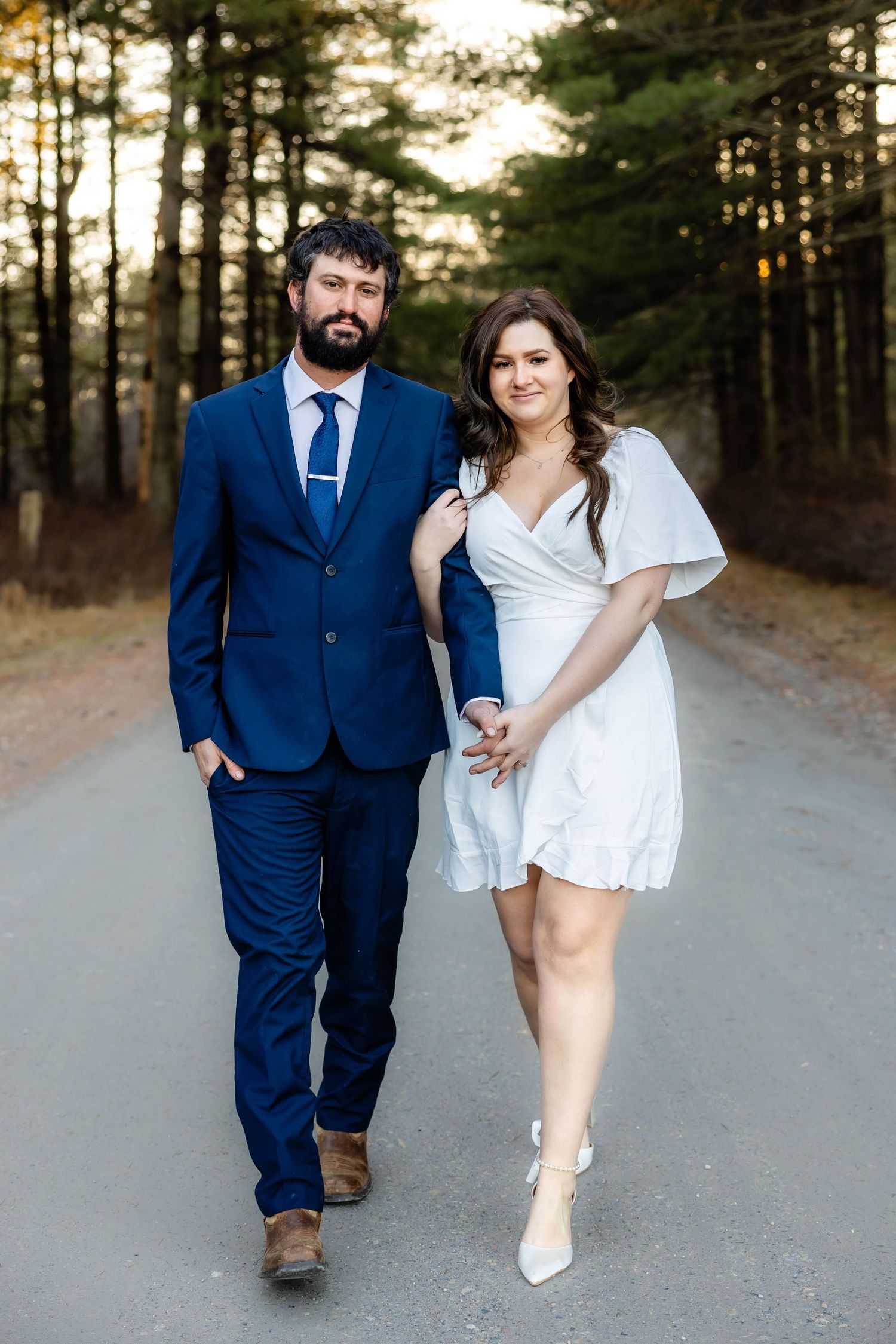 A couple poses together on a dirt road surrounded by pine trees during golden hour.