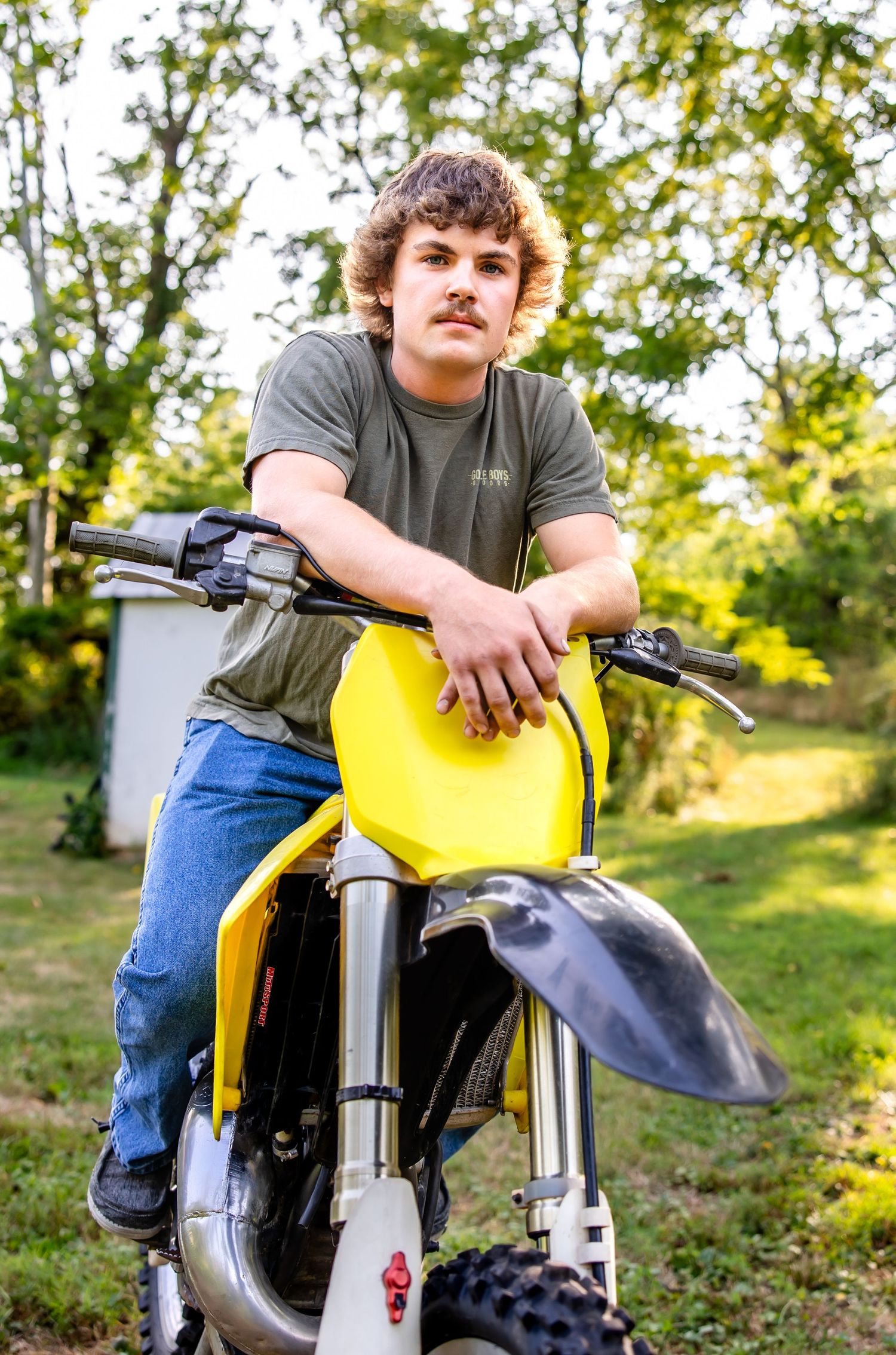 Person poses with yellow dirt bike in wooded area during outdoor photoshoot.