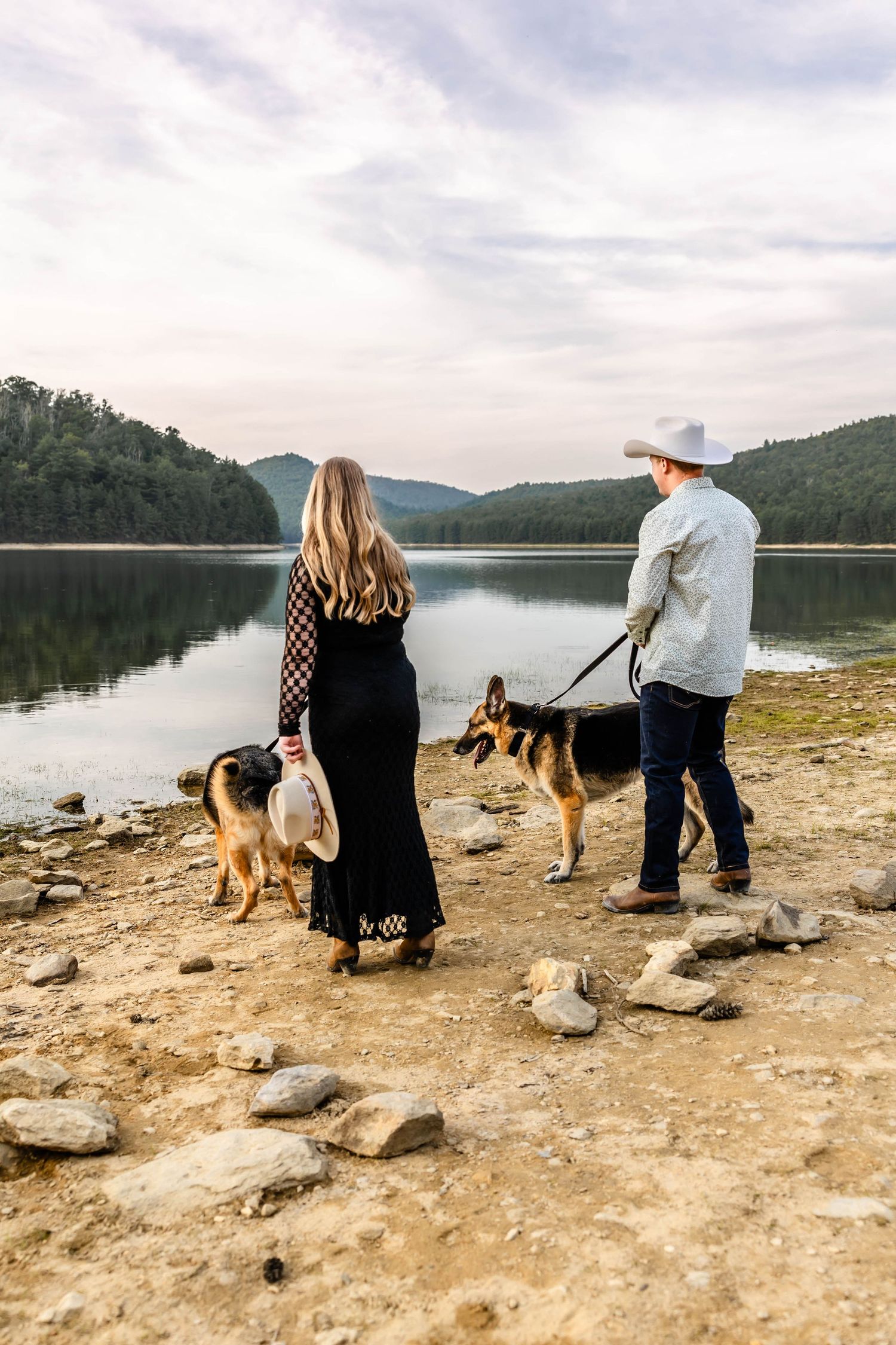 A couple in western attire stands with their dogs on a rocky shore overlooking a mountain lake.