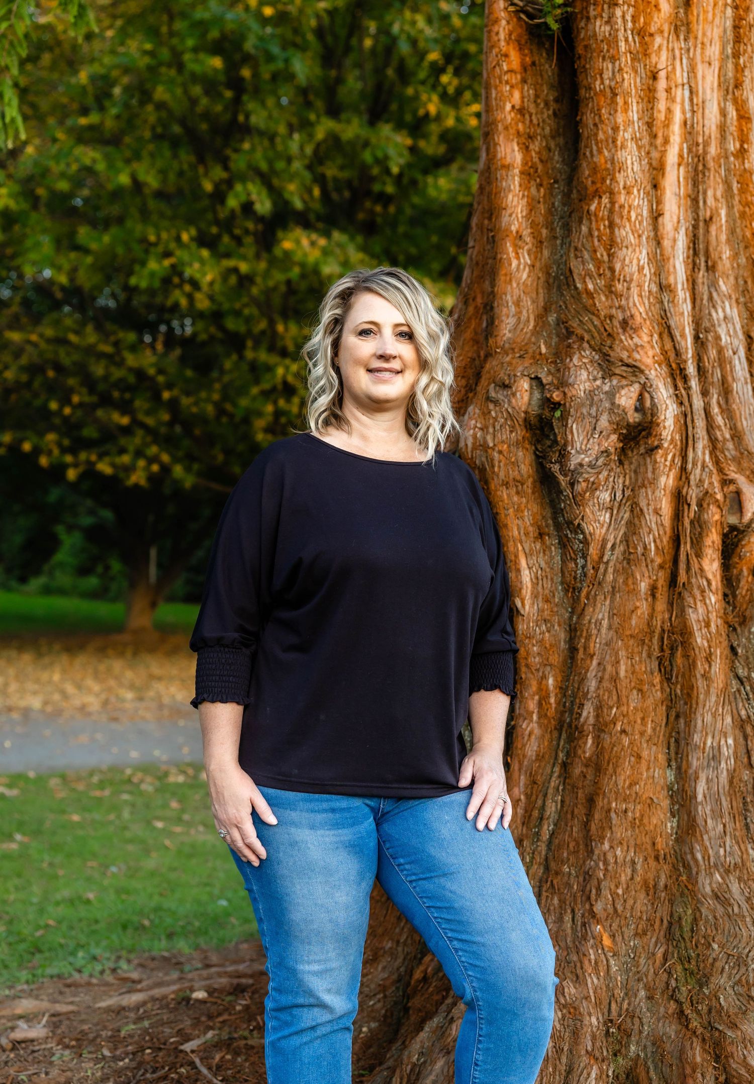 Fashion portrait of a person in a black sweater and blue jeans posing against a large tree trunk in an autumn park.