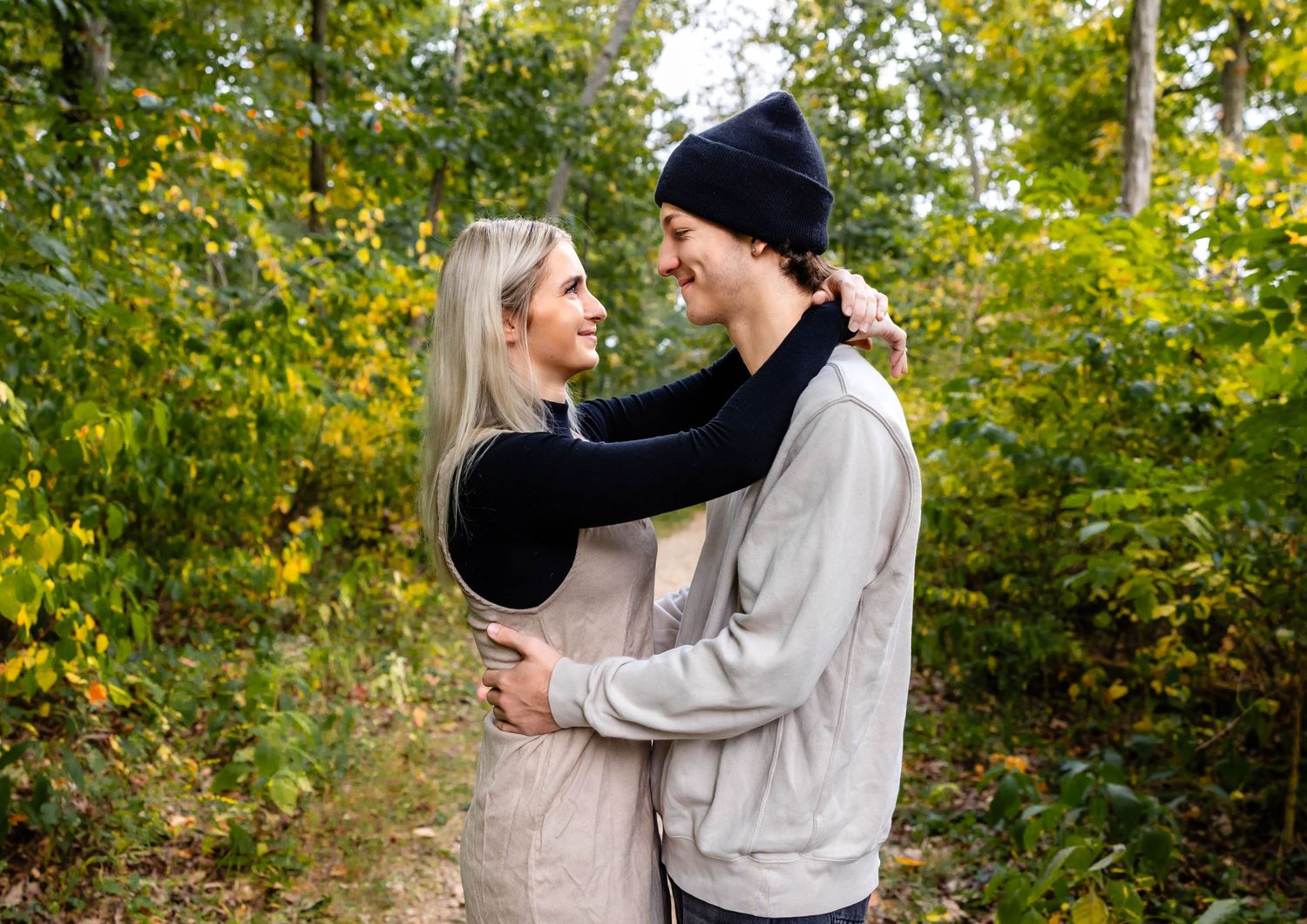 A loving couple poses together on a wooded path with autumn leaves in the background.