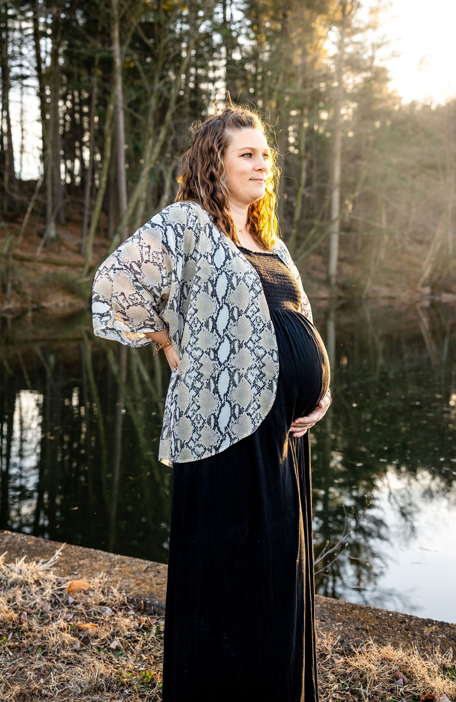 Pregnant woman in snake print kimono poses by lake during golden hour.