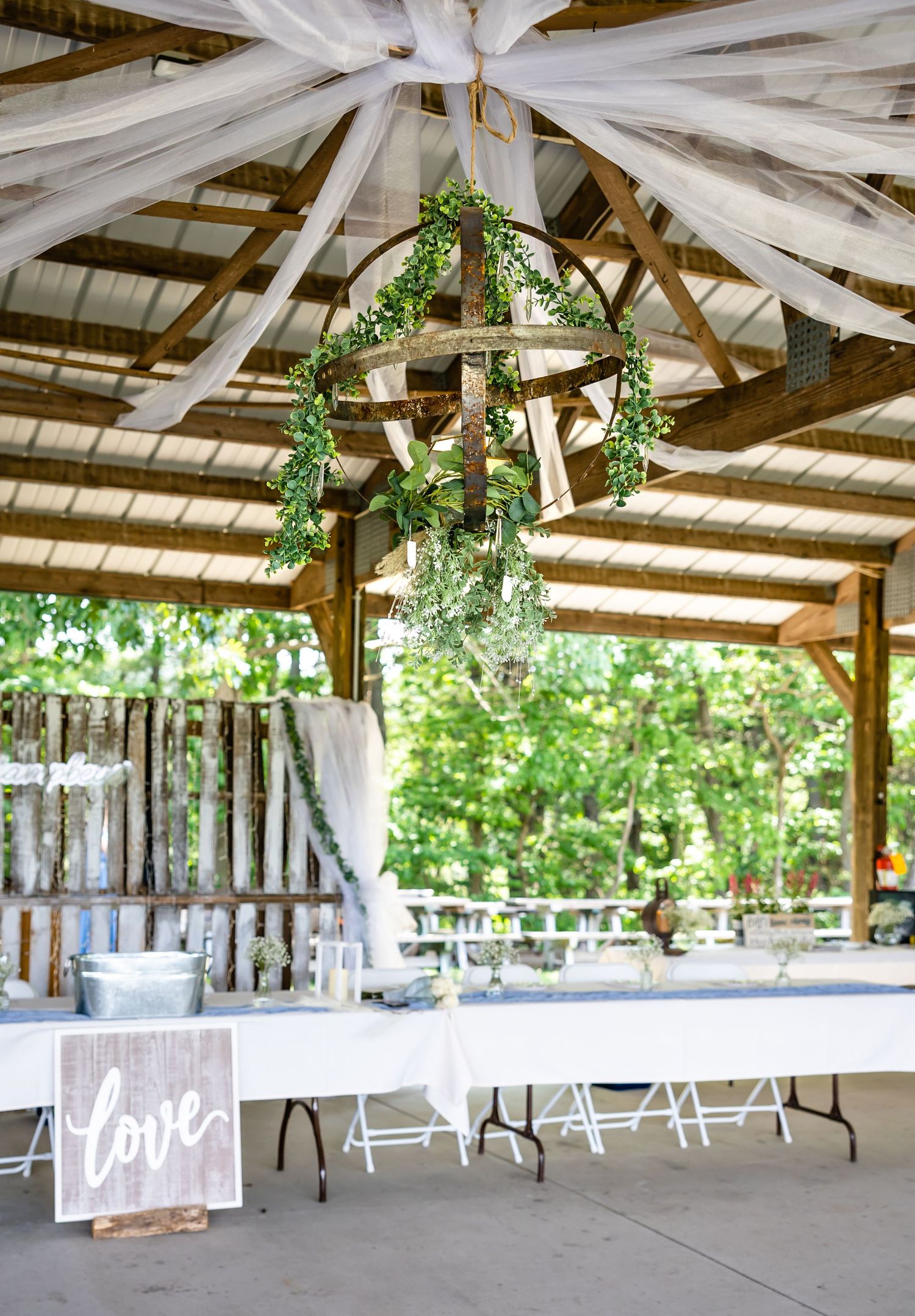 Rustic outdoor wedding pavilion decorated with white fabric draping and greenery hanging from wooden beams.