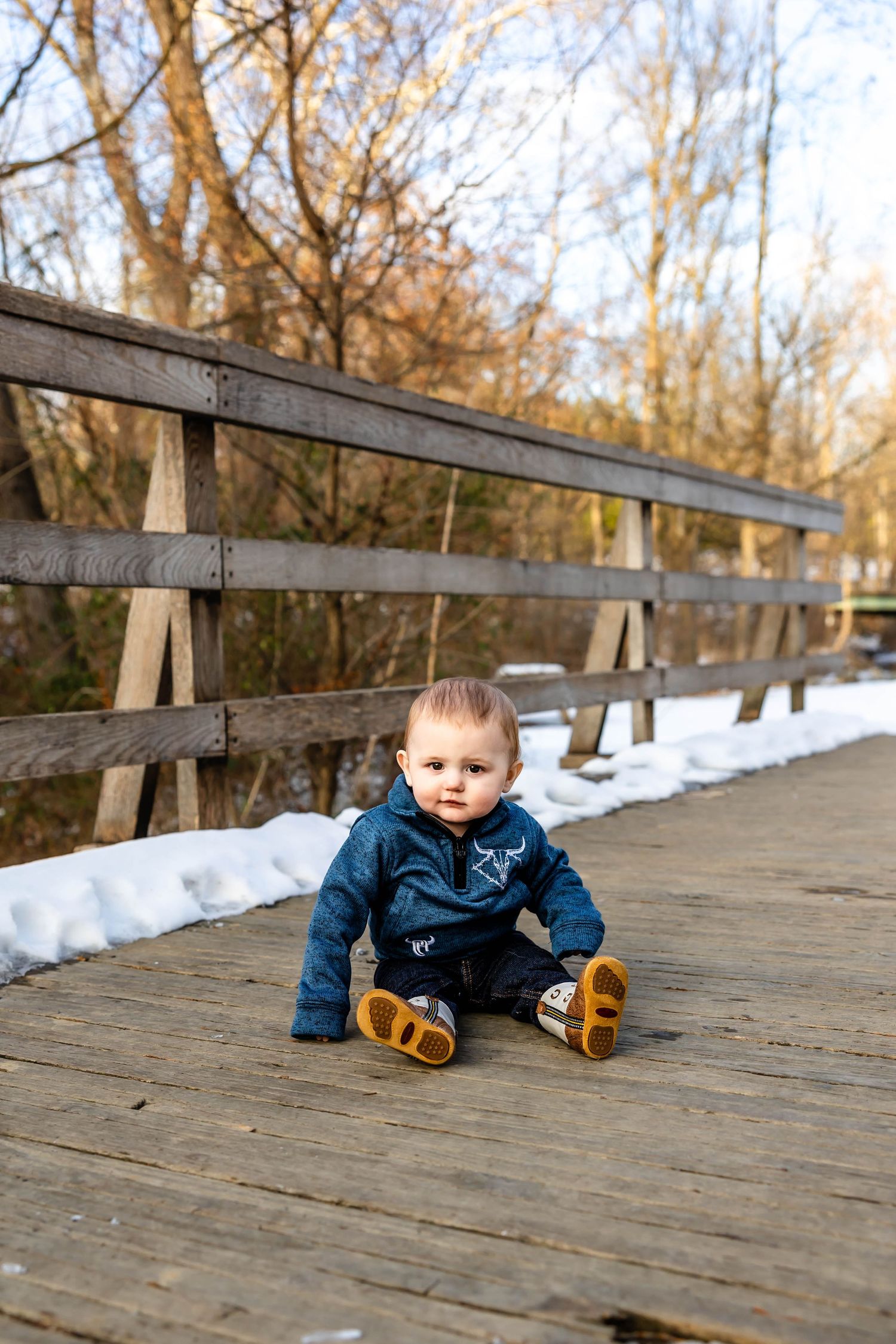 A small child in a blue sweater sits on a wooden boardwalk surrounded by snow.