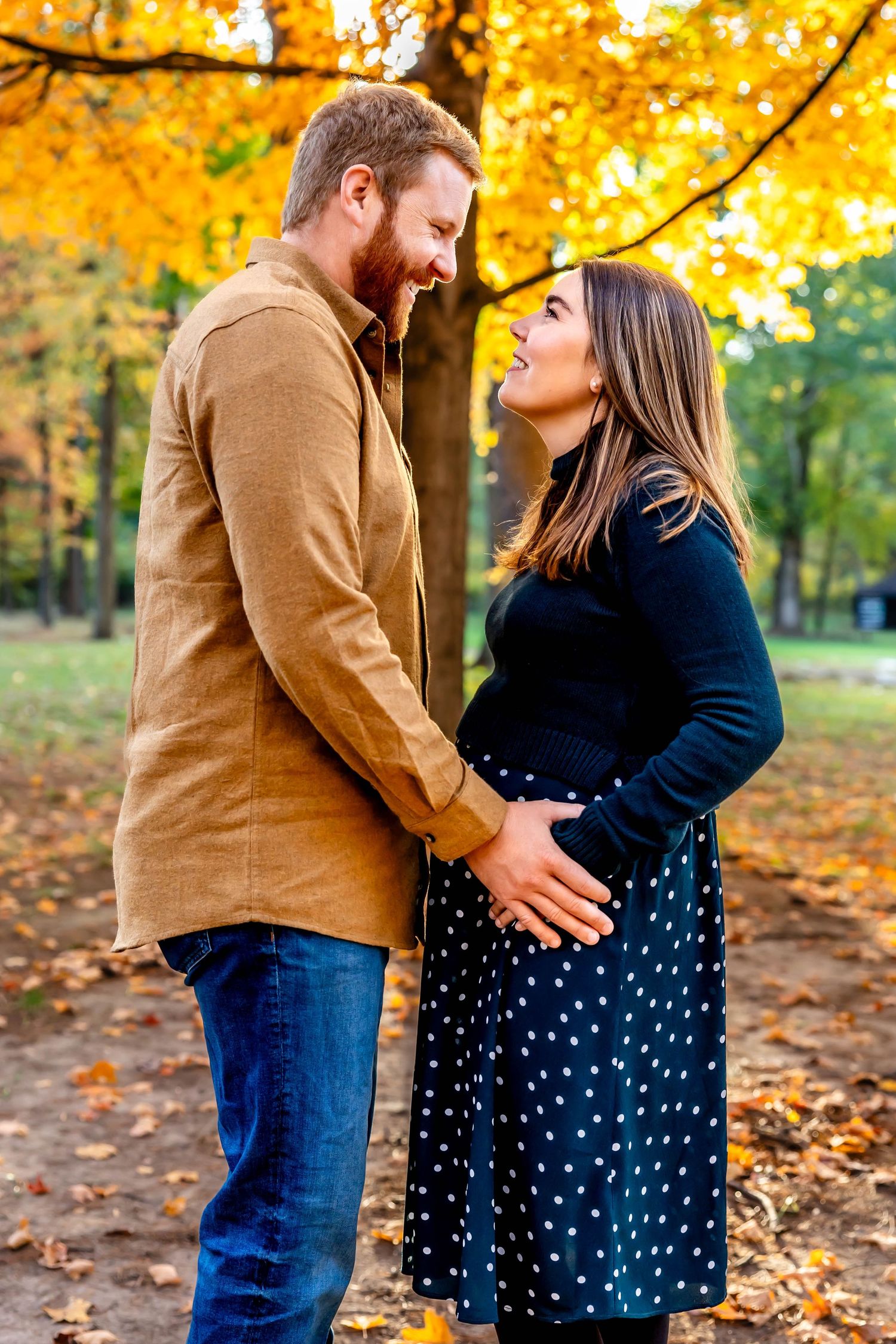A couple shares an intimate moment in a park surrounded by golden autumn foliage.