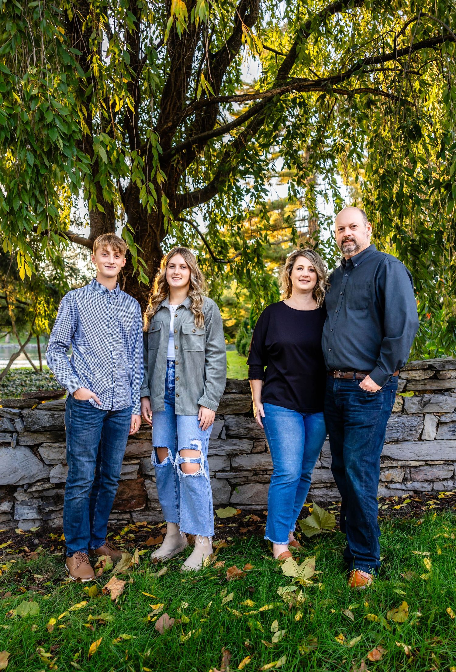 Group standing by stone wall with autumn trees and yellow leaves in a casual outdoor setting.
