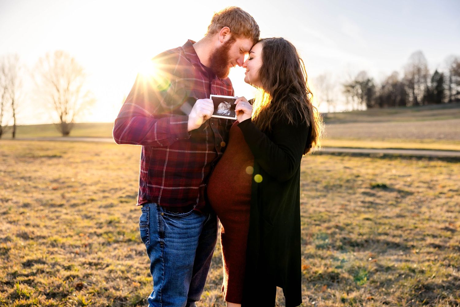 Couple sharing a tender moment at sunset in an open field while holding a sonogram photo.