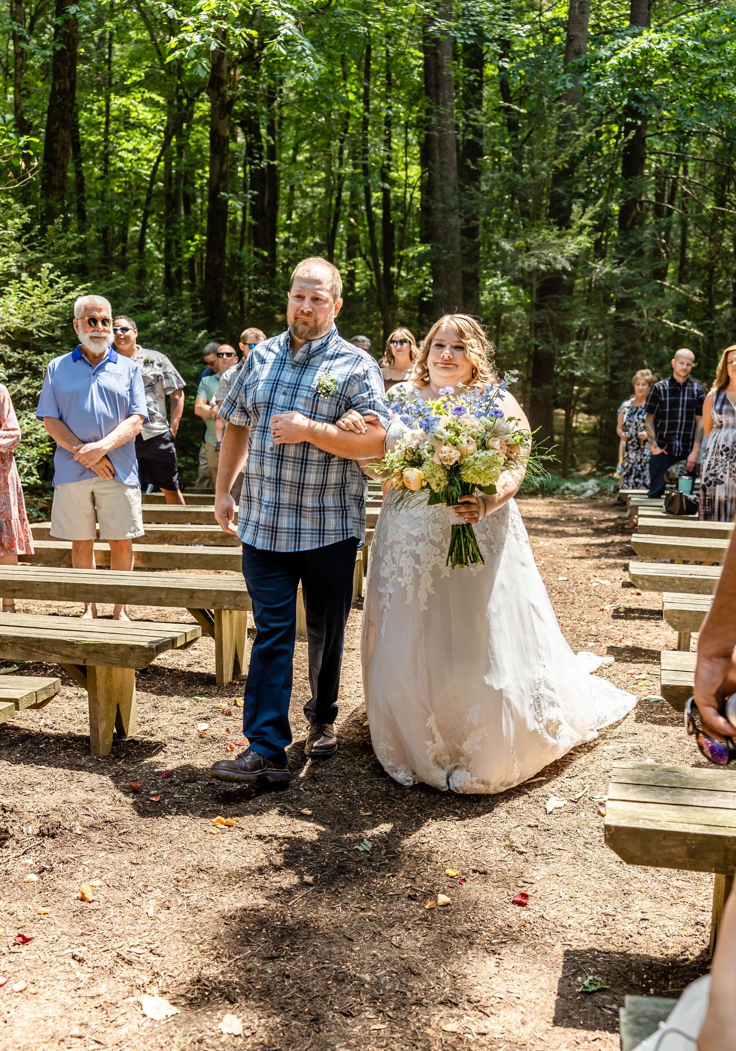 Outdoor forest wedding ceremony with wooden benches and guests gathered on a dirt path.
