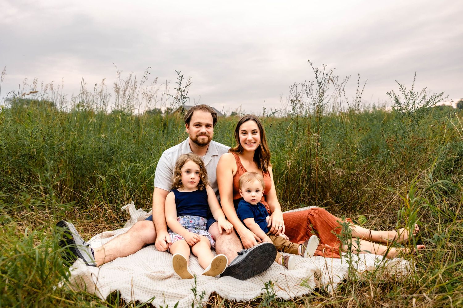 Family relaxing together on a white blanket in a wild grass field during summer.