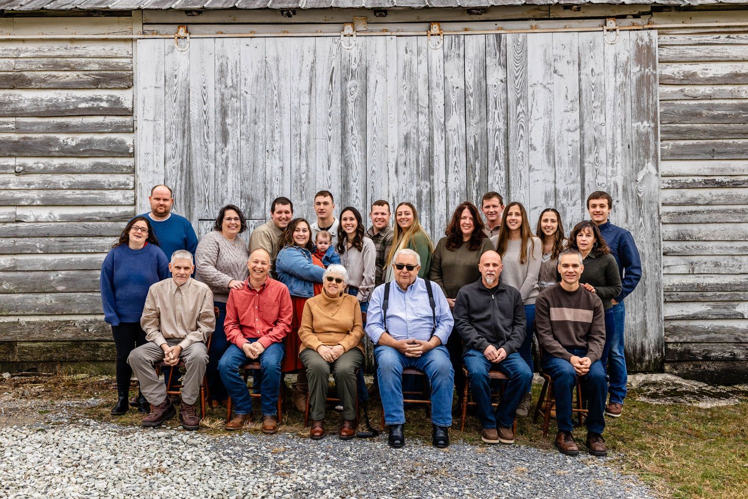 Large group portrait of people seated in rows against weathered wooden barn wall.