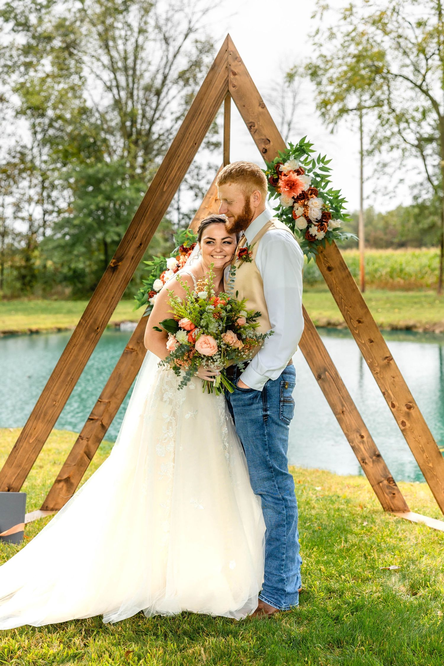 A couple embraces under a rustic wooden triangle arch during their outdoor wedding ceremony.