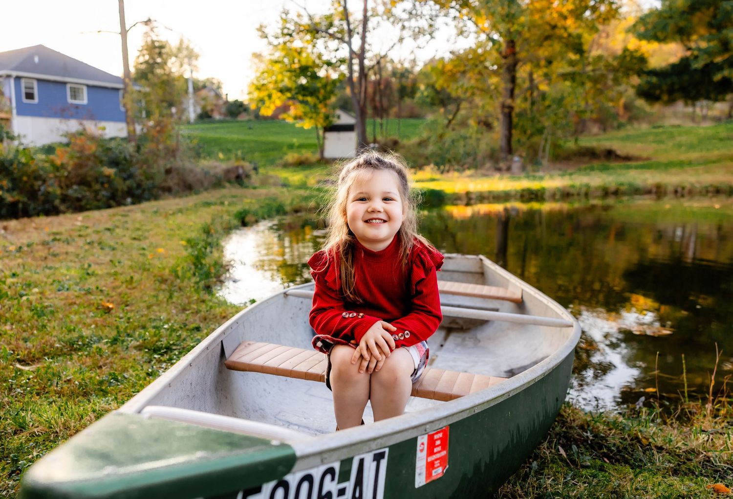 A happy child in red sits in a numbered green canoe during autumn by a blue house.