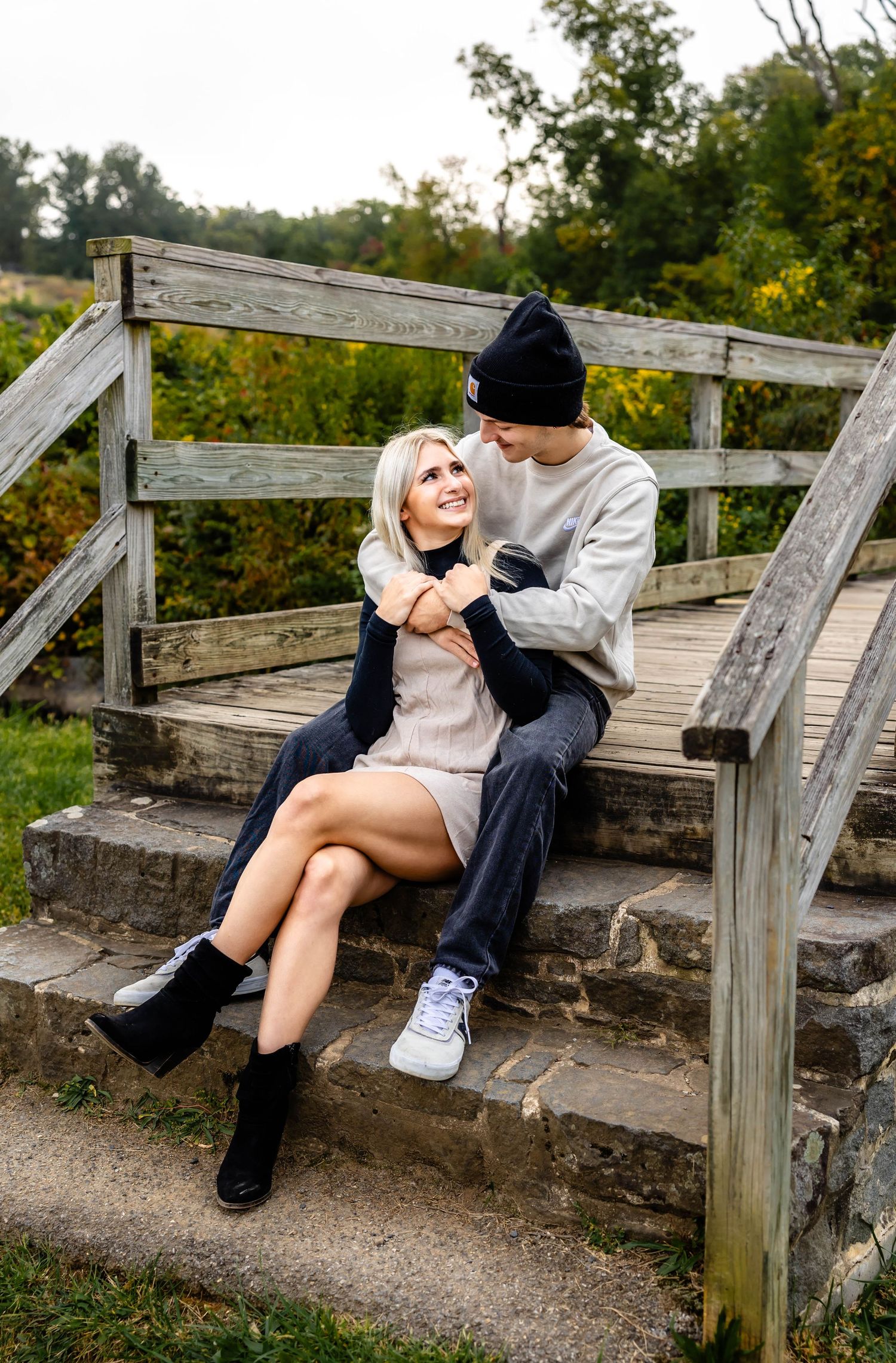 A couple embraces on wooden steps with autumn trees in the background.