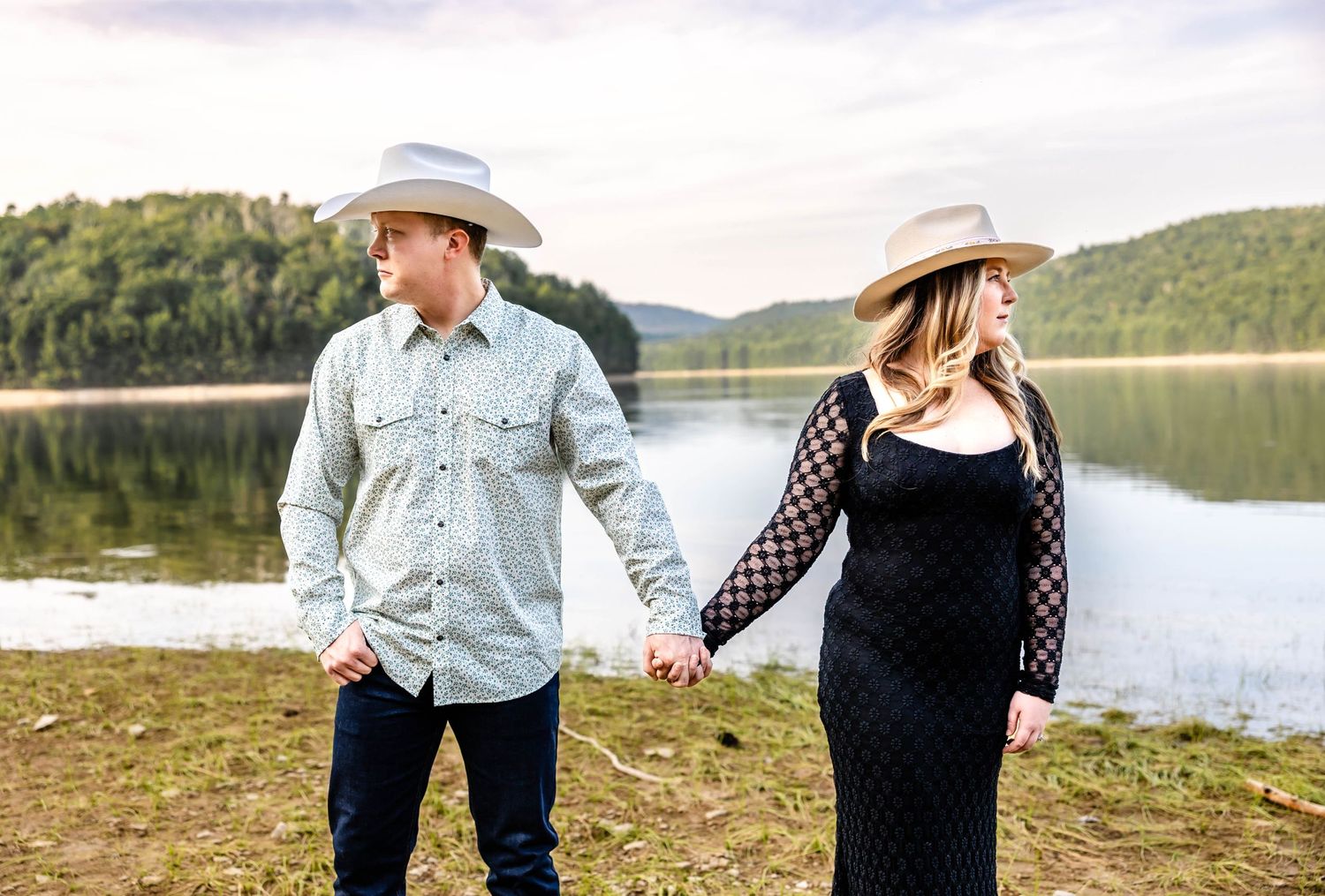 Couple in western wear walks along lakeside holding hands during sunset.