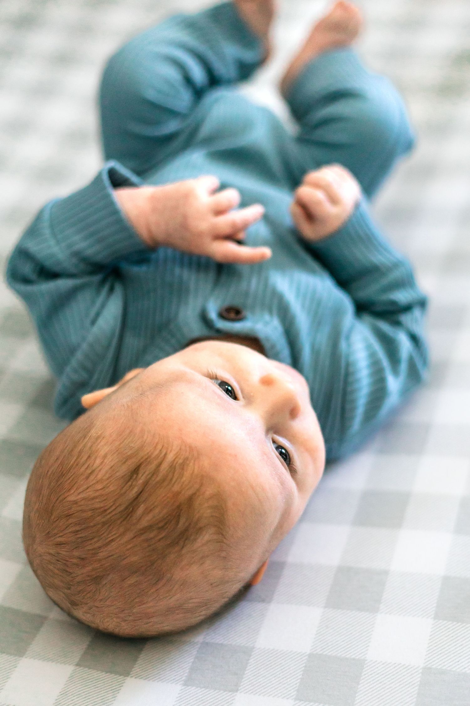 Baby in teal ribbed outfit lying on checkered blanket looking up at camera.