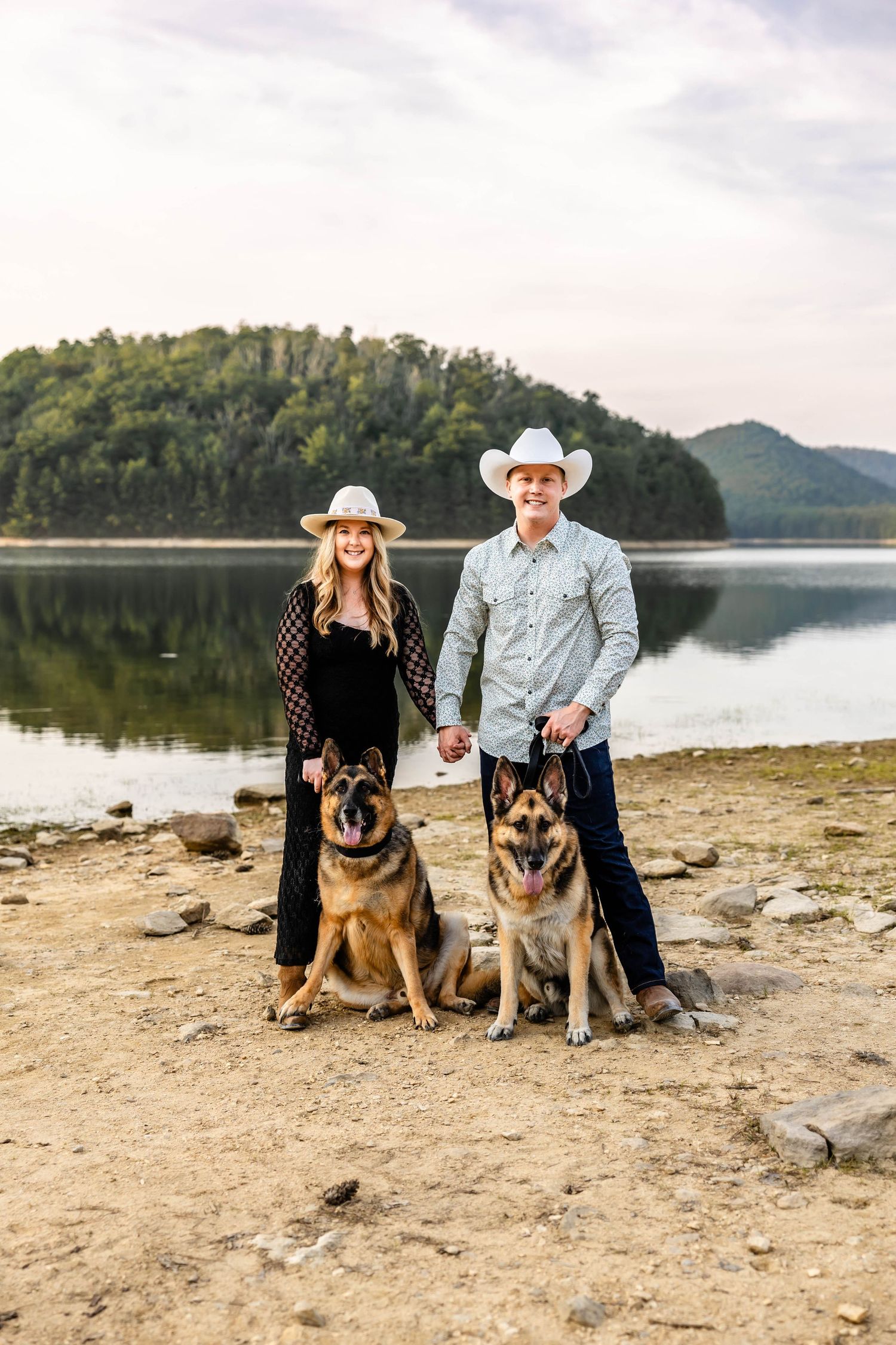 A couple in western wear poses with their two German Shepherd dogs by a serene lake.