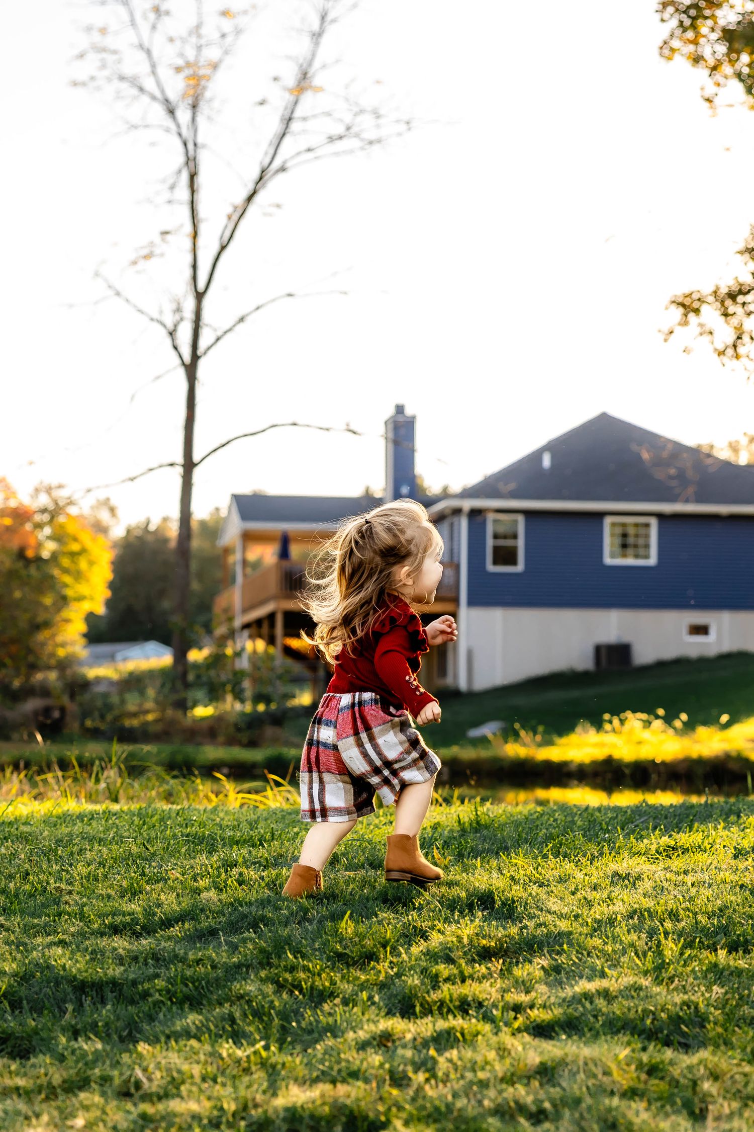Child in red sweater and plaid skirt dances in backyard with blue house visible in background.