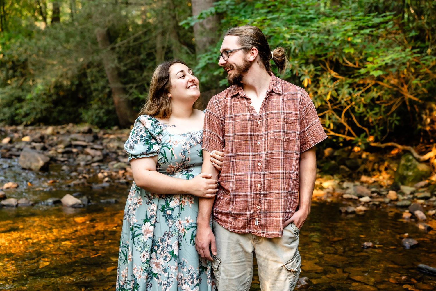 A couple laughs together while standing by a creek in a lush forest setting.