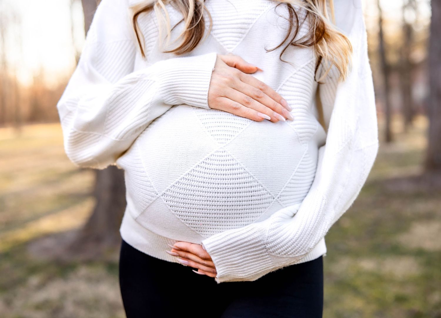 Close up of pregnant belly in textured white sweater against sunset background.