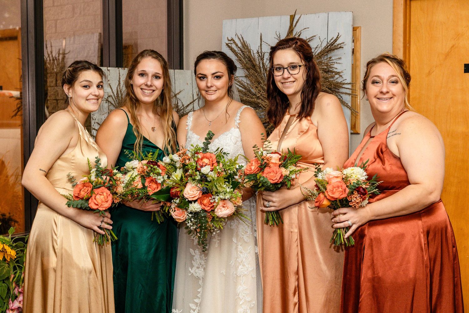 Bridesmaids in jewel-toned dresses pose with their bouquets at indoor venue.