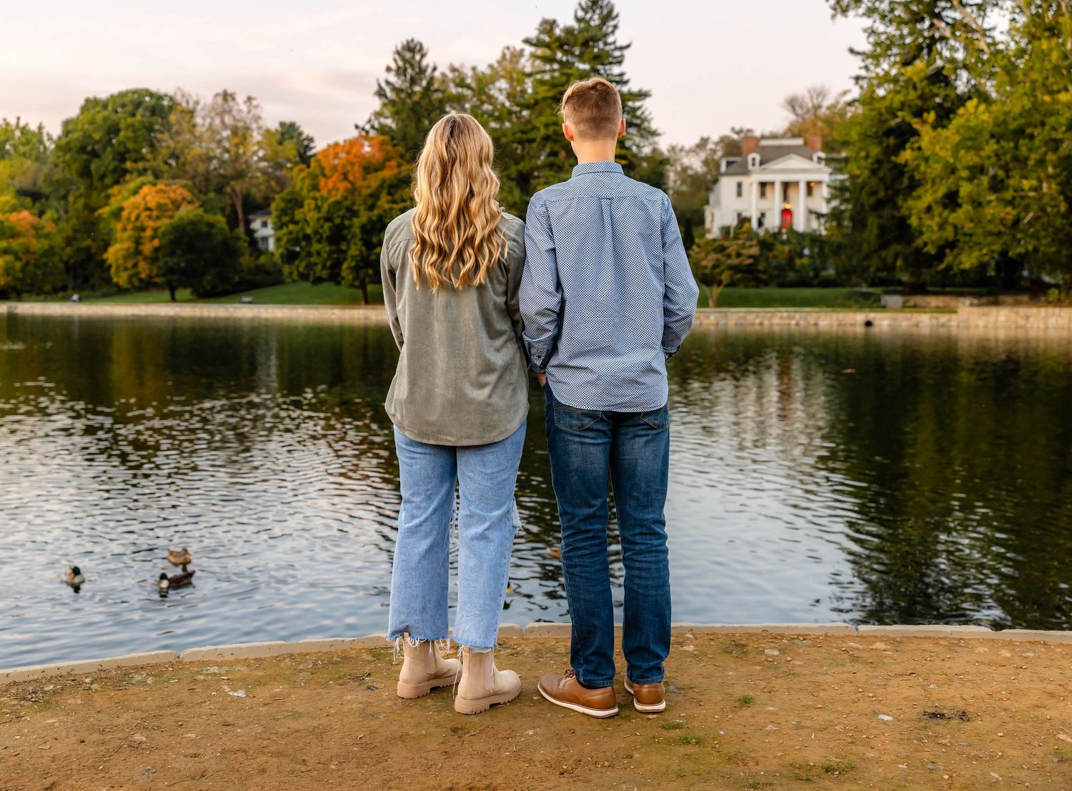 Couple standing together looking at a lake during autumn with trees and a house in background.