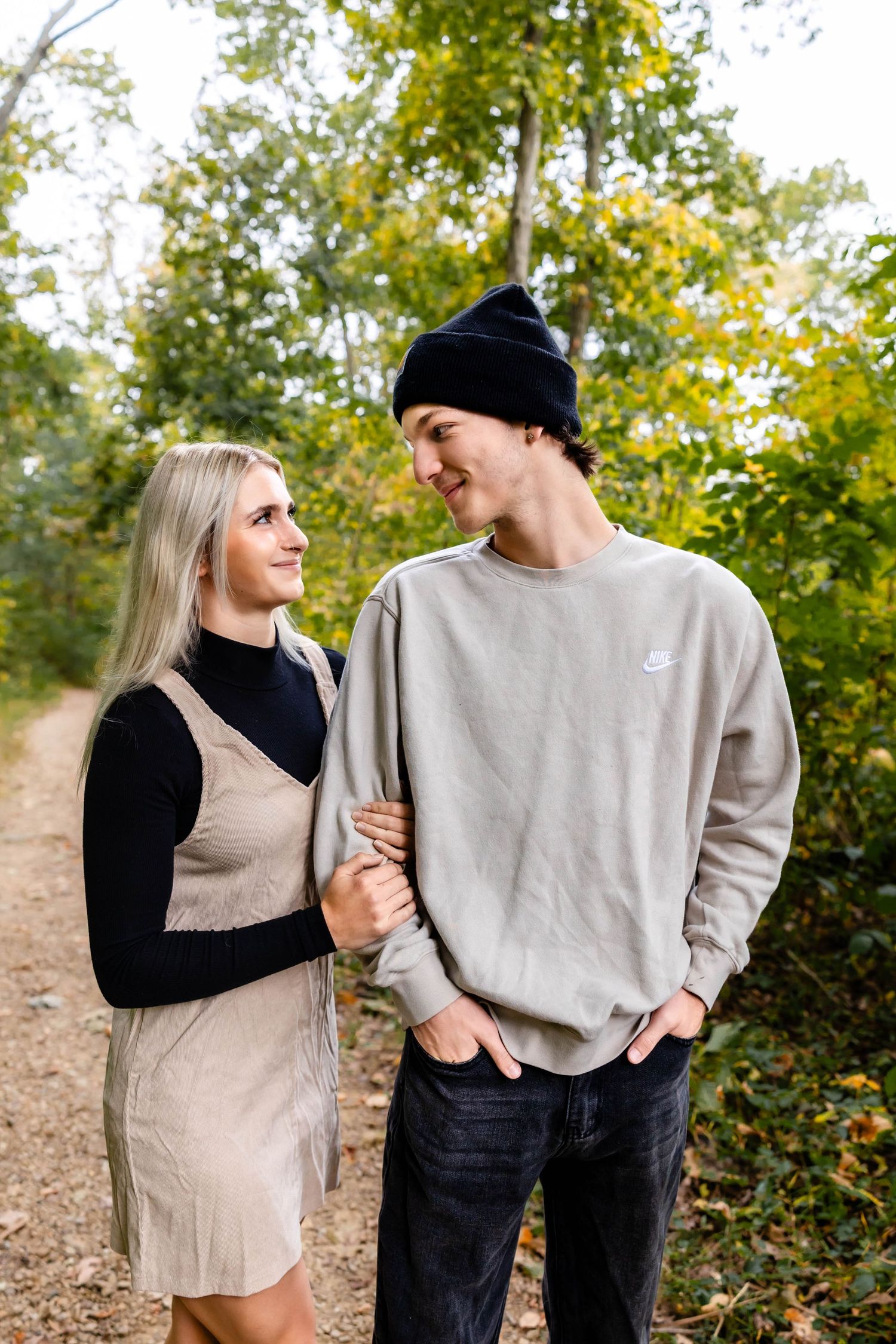 A couple gazes at each other lovingly while standing on a fall trail with trees in the background.