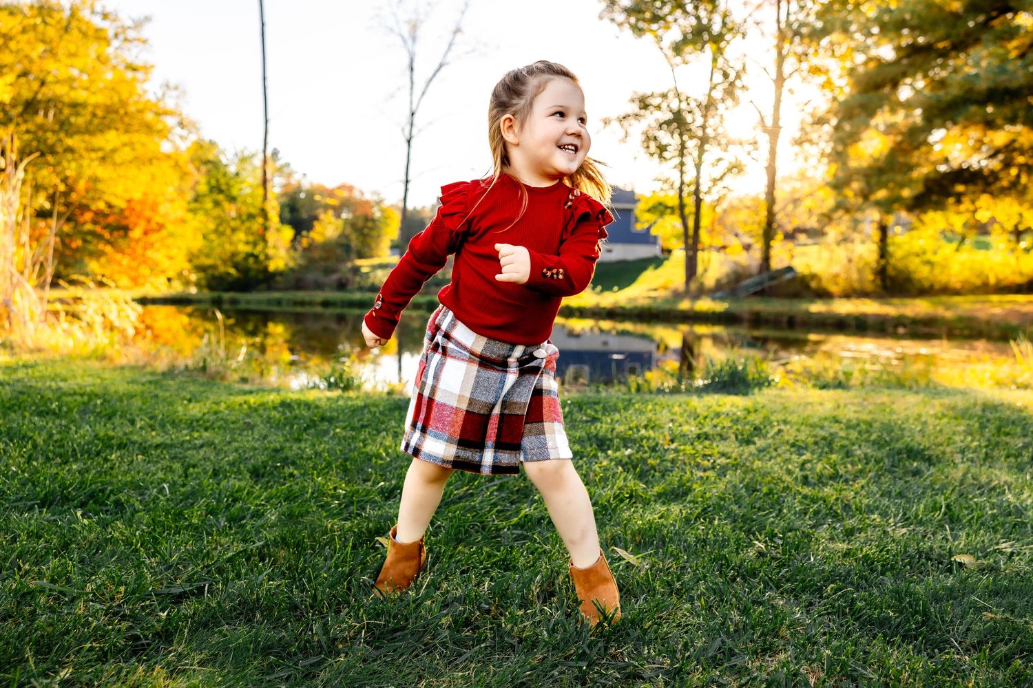 Child in festive red and plaid outfit plays outdoors against autumn trees and pond.