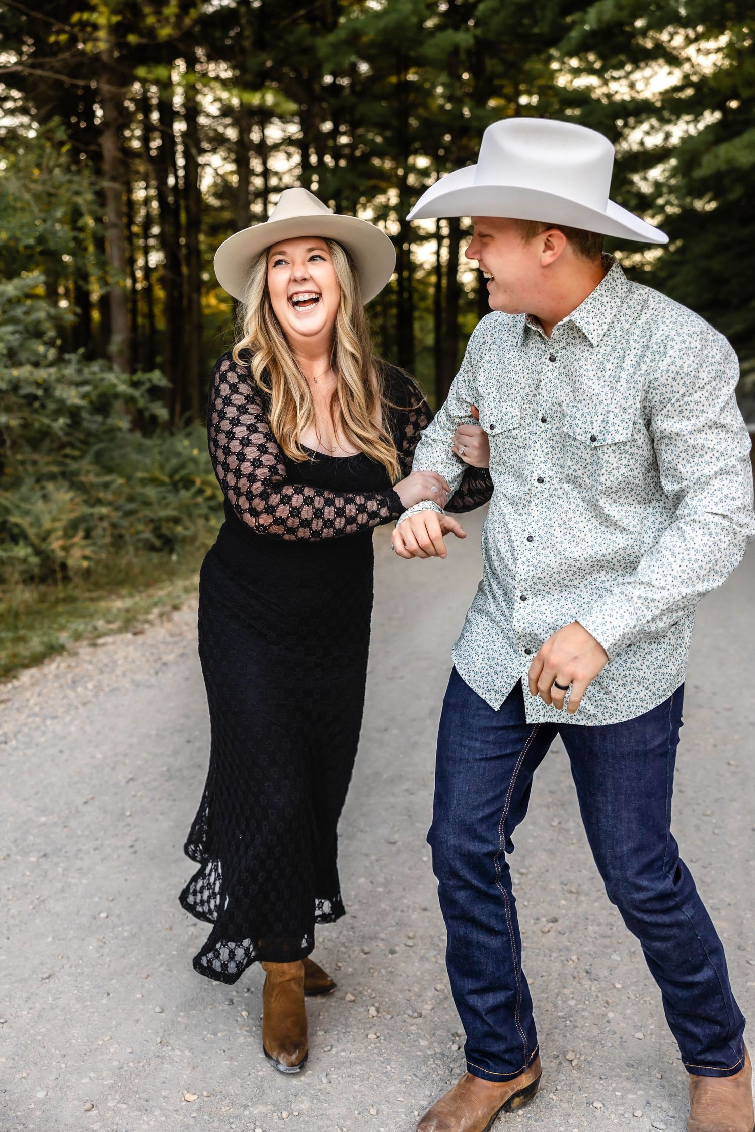 A couple in cowboy hats dancing and laughing together on a dirt road surrounded by pine trees.