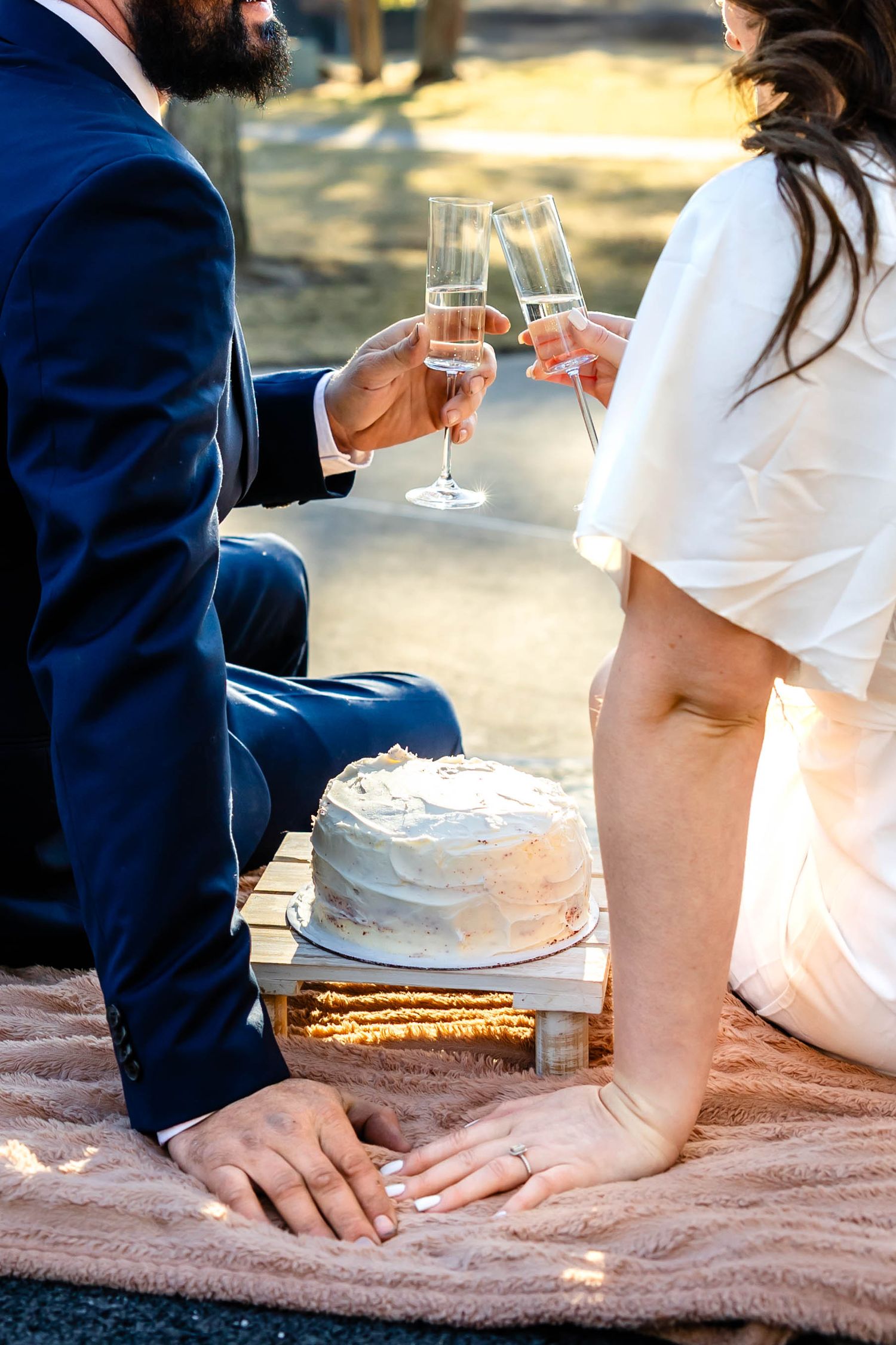 Two people toast with champagne glasses beside a white cake on a blanket.