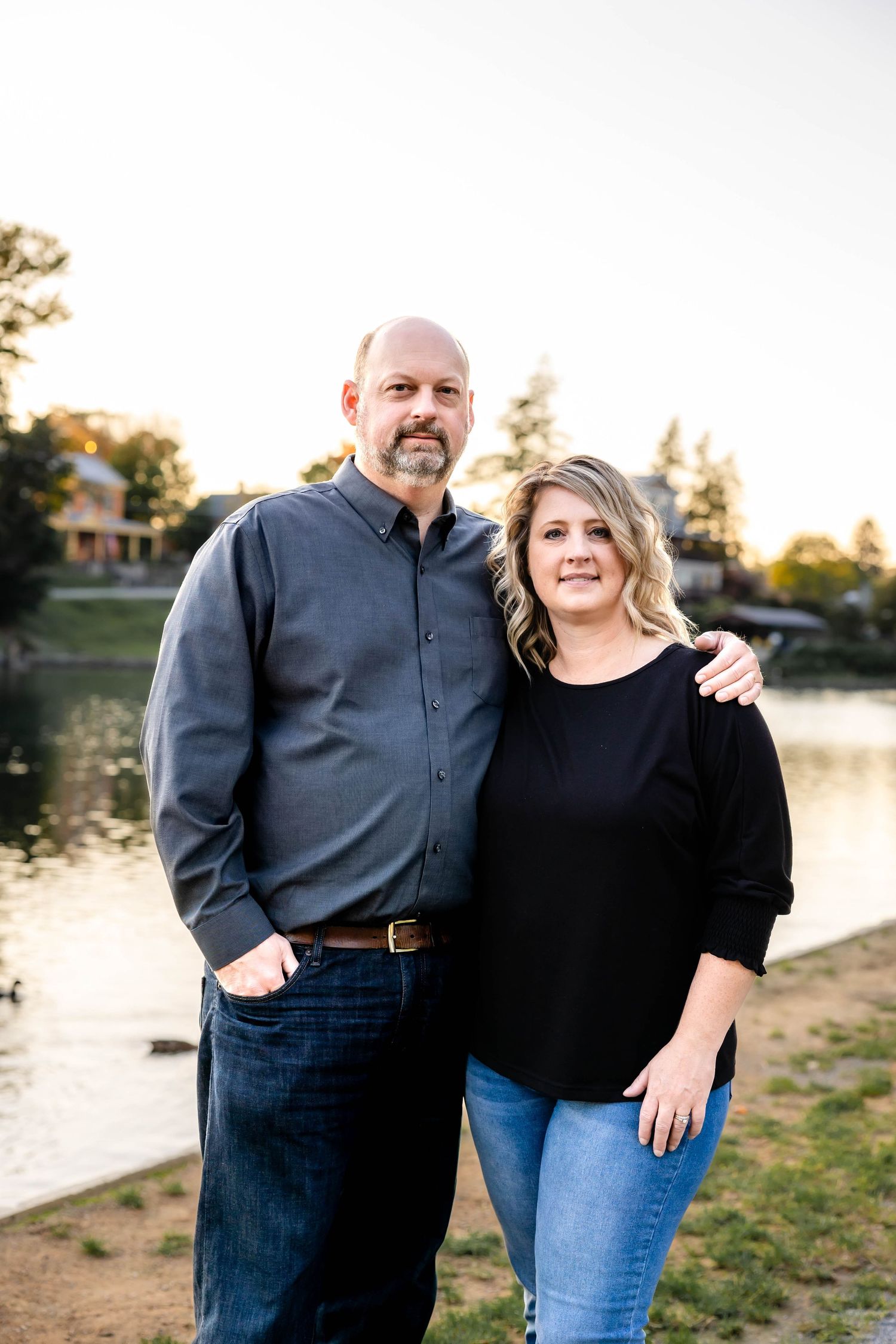 A couple poses together by a lake during sunset wearing casual attire.