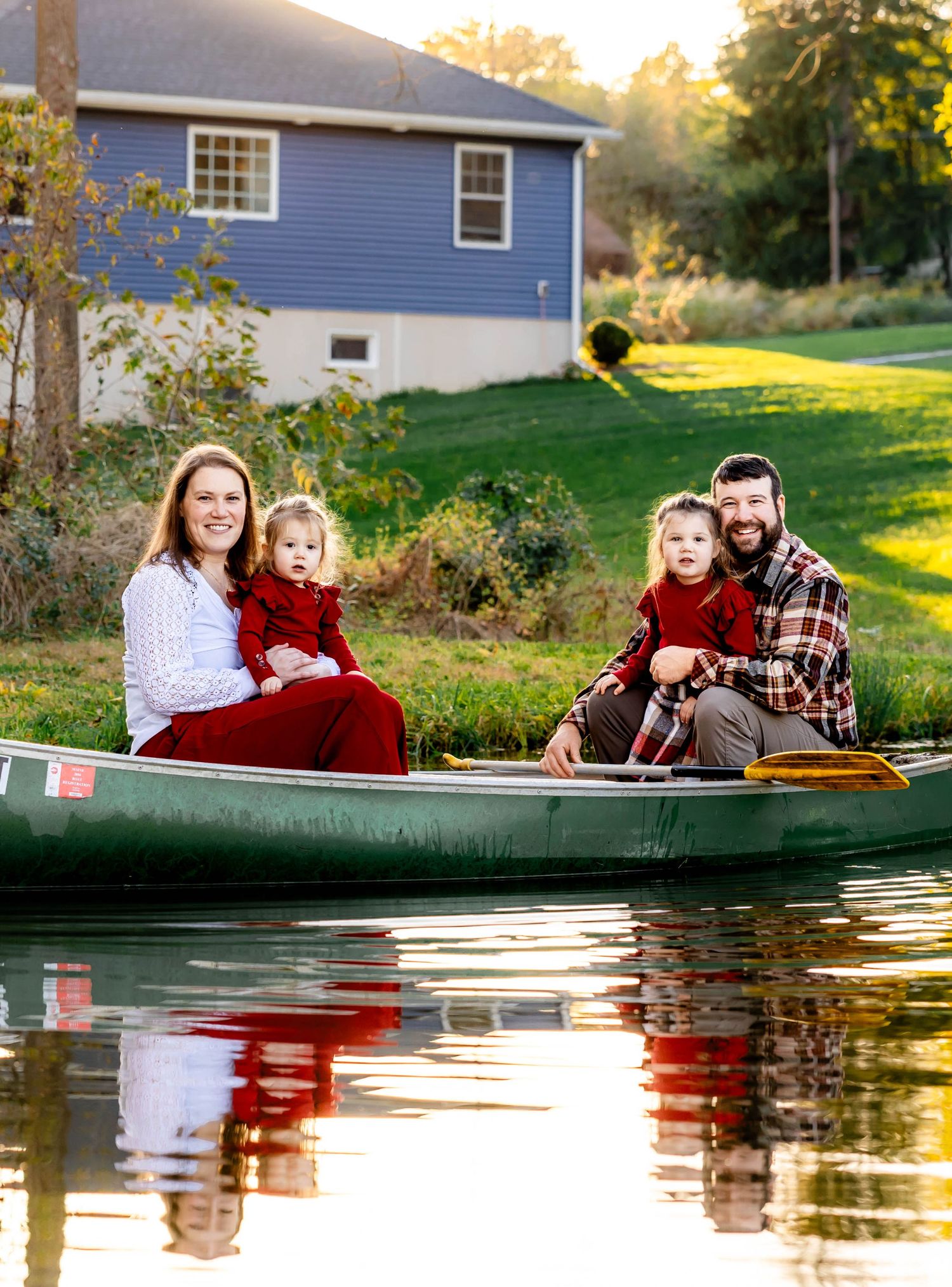 A family sits in a green canoe on a tranquil pond beside a blue house during golden hour sunset.
