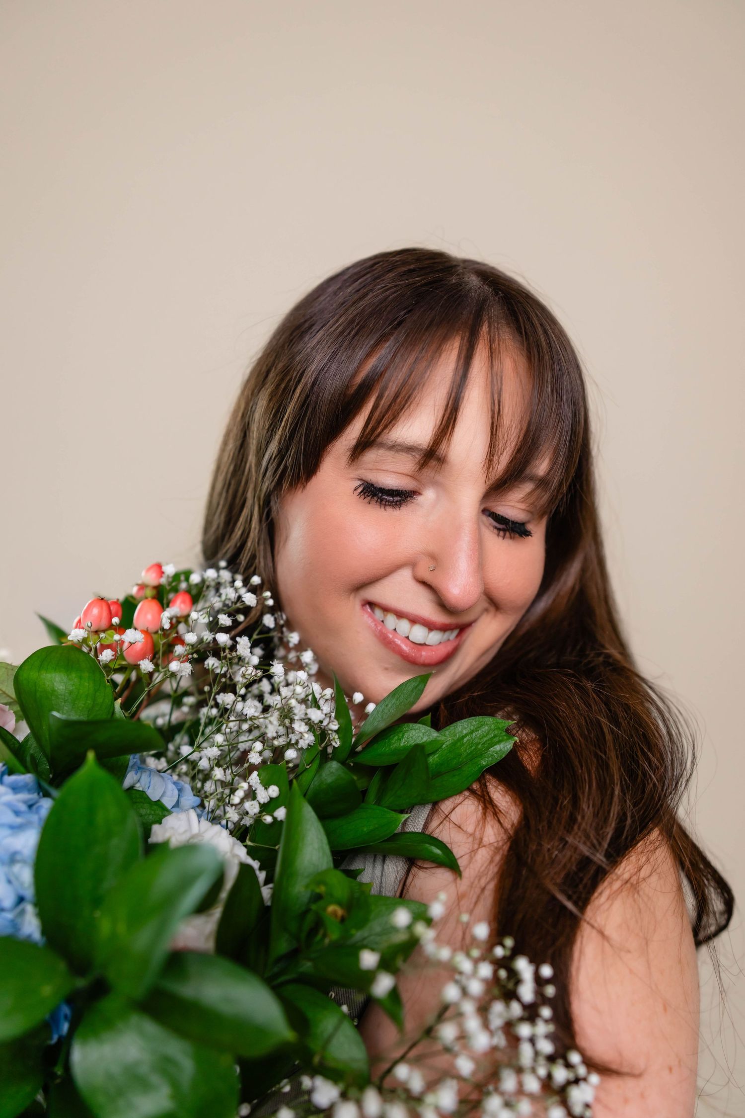 Smiling brunette woman with bangs holding a bouquet of pink roses, blue hydrangeas, and baby's breath.