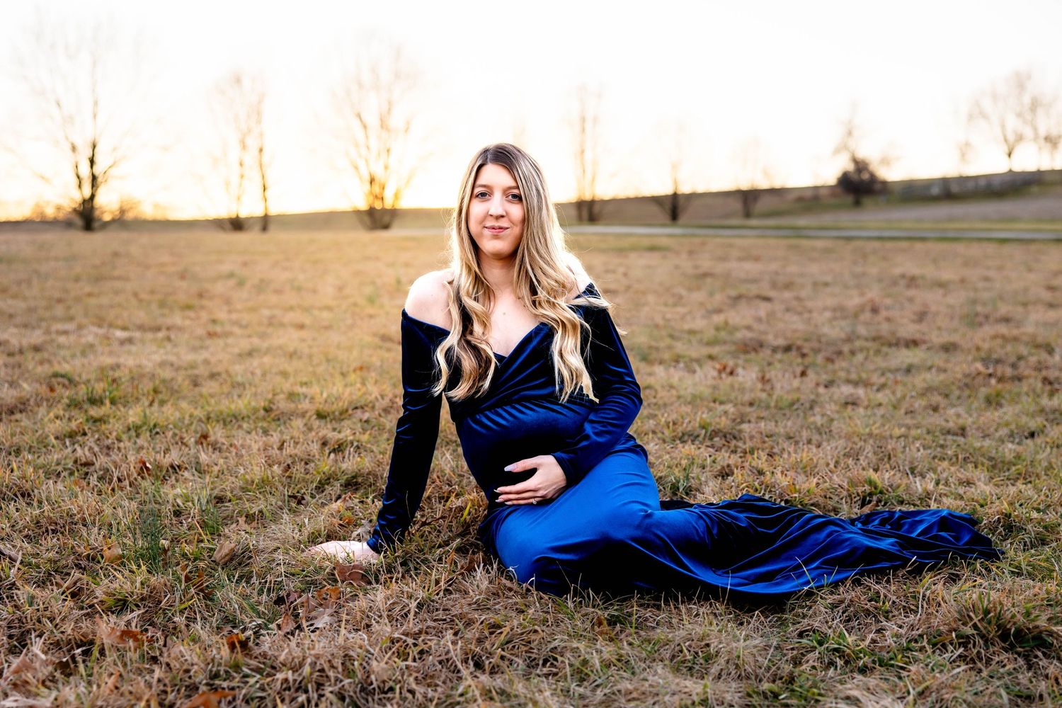 Elegant maternity photo with woman in navy dress sitting in golden field at dusk.