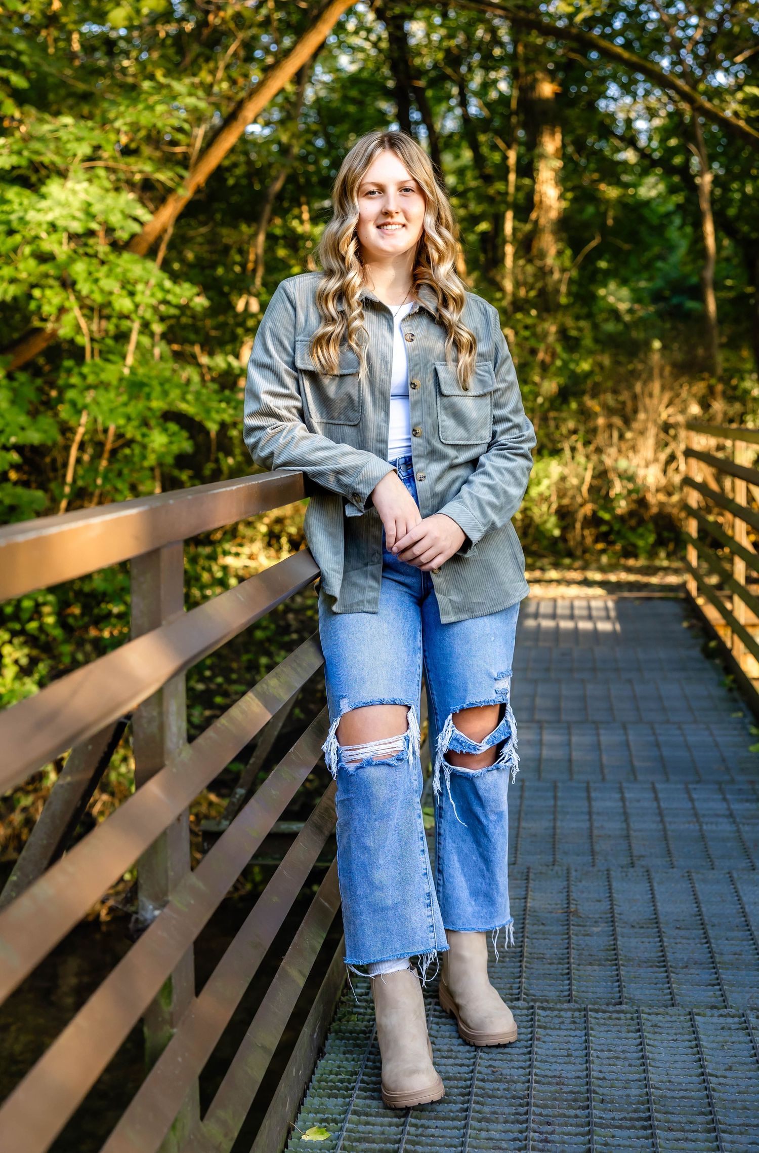 Young woman in distressed jeans and gray jacket stands on wooden walkway in park setting.