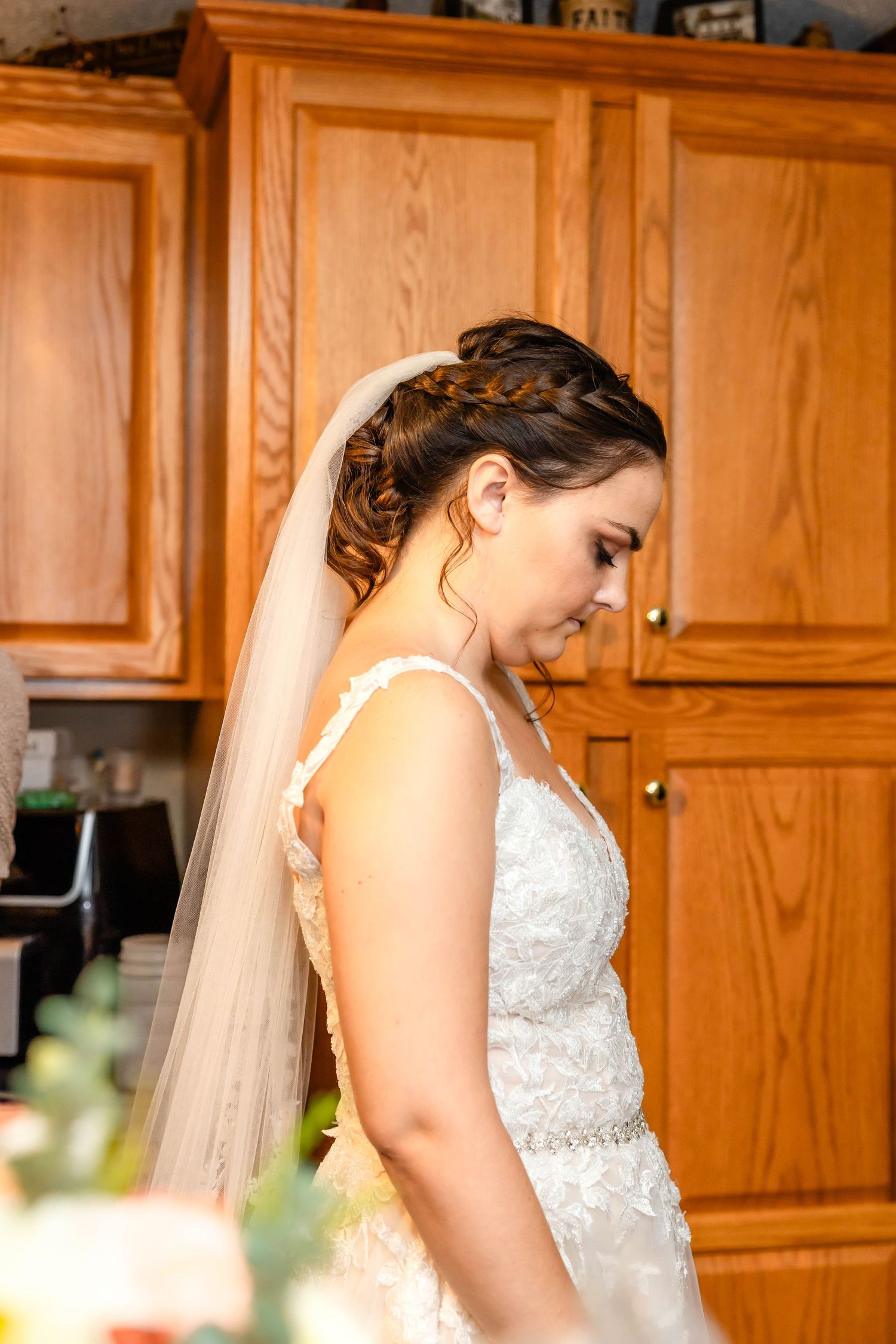 A bride in a white lace wedding dress stands in front of wooden kitchen cabinets.