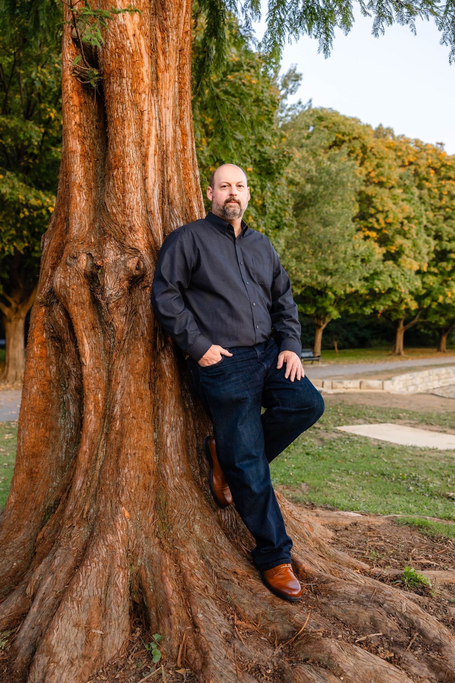 Person in dark jacket and jeans poses confidently against massive tree trunk in park setting.