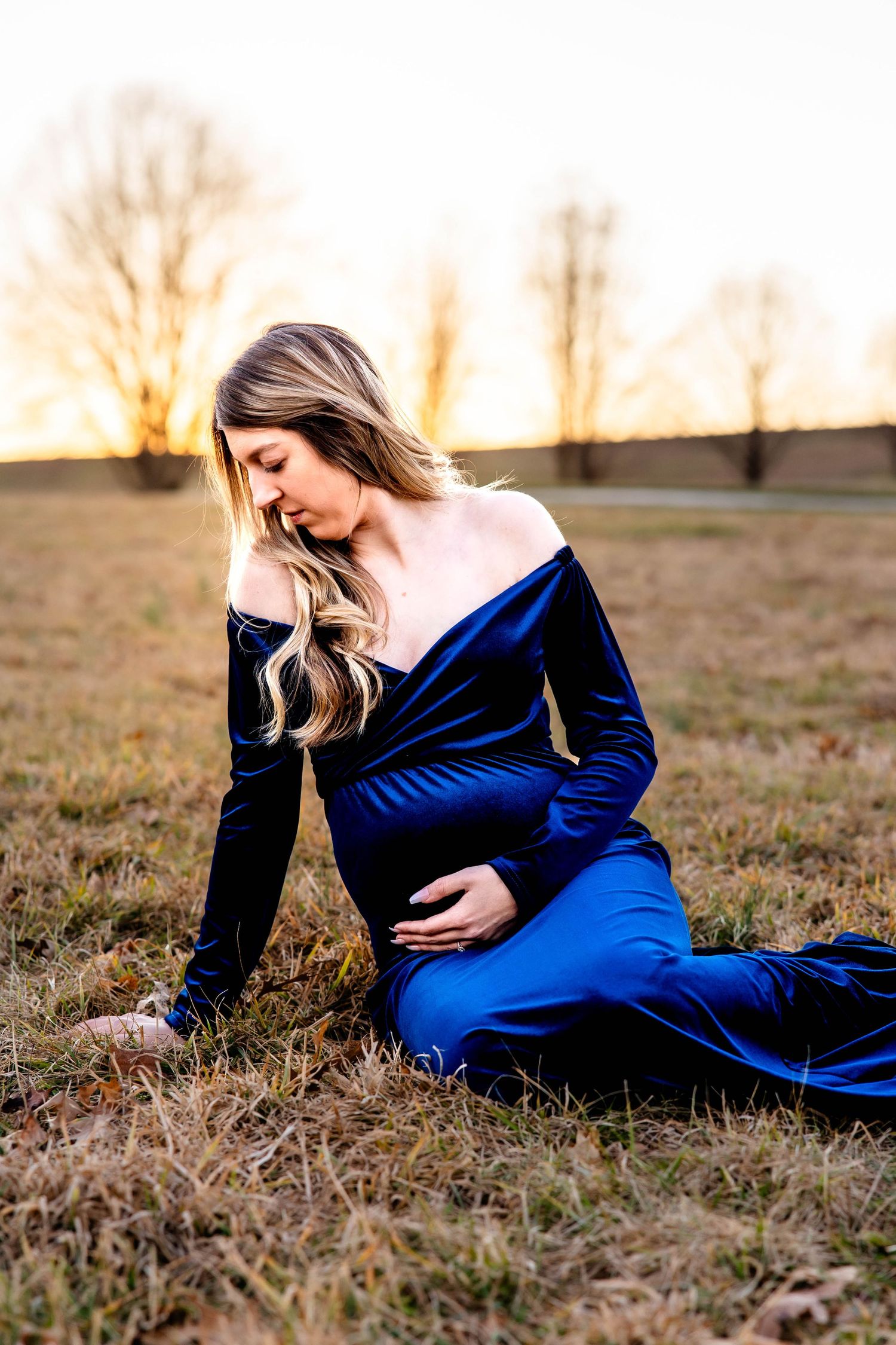 A person wearing a deep blue velvet gown sits thoughtfully in a golden field during sunset.